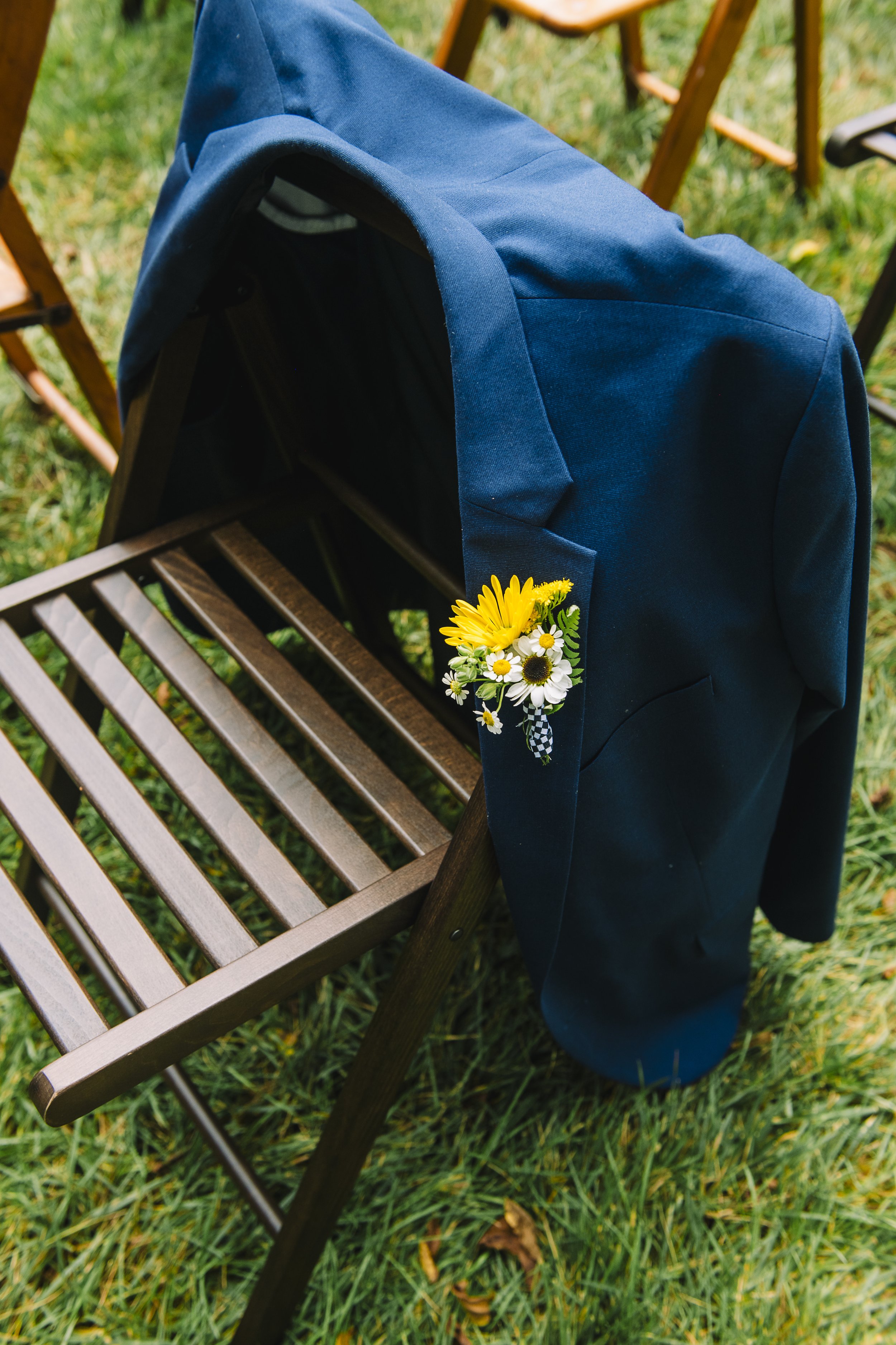 A blue jacket with a small bouquet of yellow and white flowers pinned to the sleeve, draped over a wooden chair on a grassy outdoor setting.