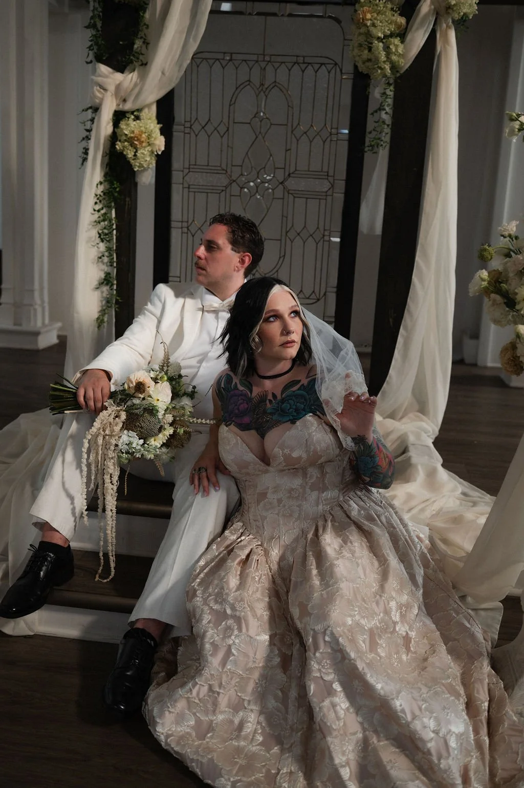 A man and a woman dressed in wedding attire sitting on a bench in a decorated setting, with the woman displaying tattoos on her arms and chest, and the man holding a bouquet of flowers.