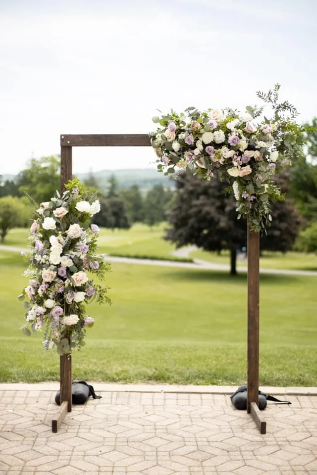 A wooden wedding arch decorated with white and purple flowers and greenery, set outdoors on a paved surface with a grassy field and trees in the background.