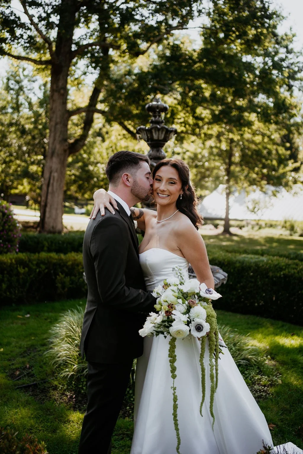 A bride and groom in wedding attire sharing a kiss outdoors, with the groom kissing the bride on the cheek, while she smiles holding a bouquet of flowers, in front of a fountain and trees.