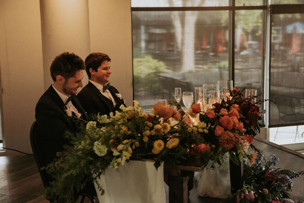Two men in tuxedos sitting at a table with floral arrangements and champagne glasses, indoors near large windows.