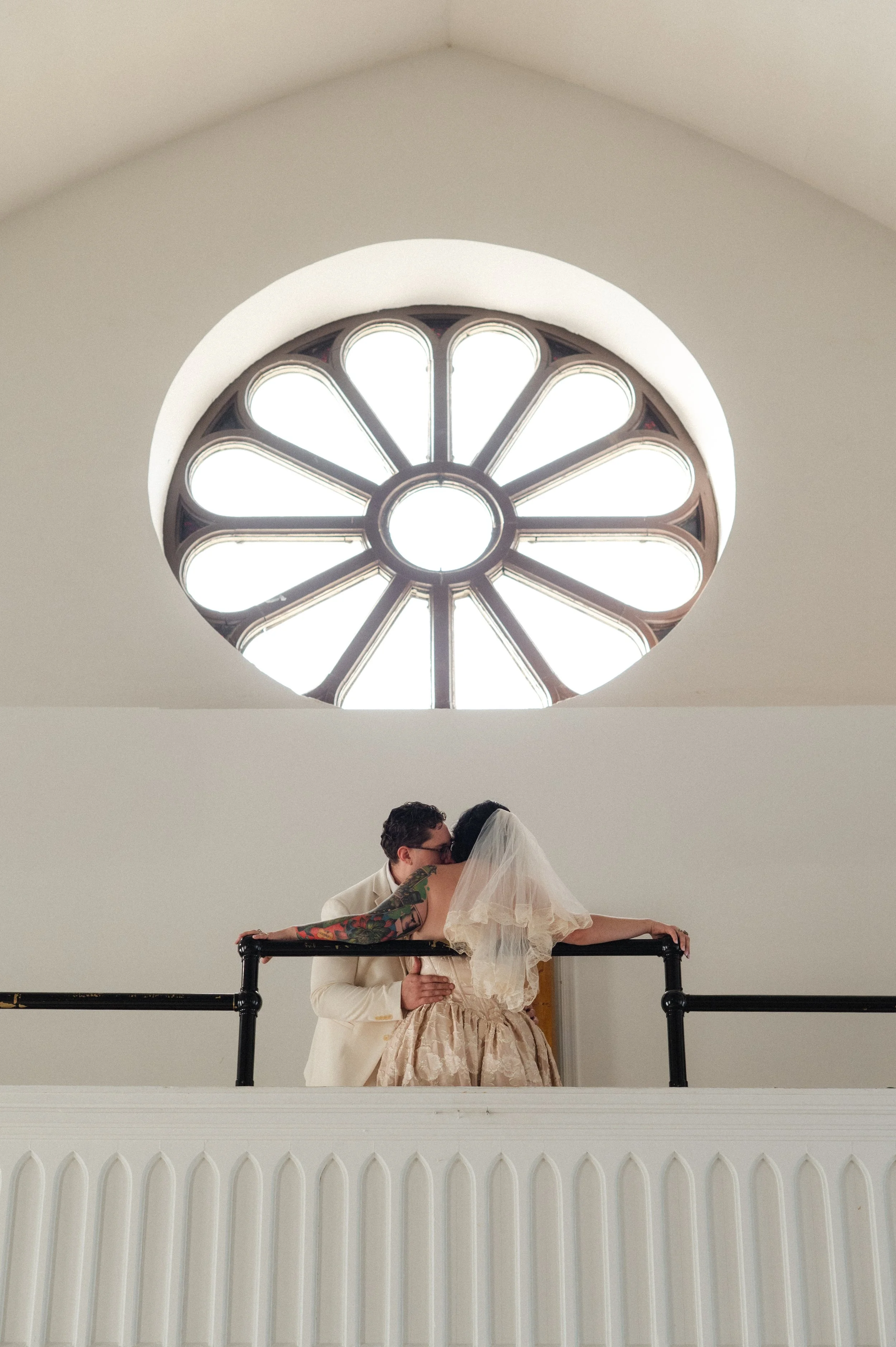 A couple kissing on an upstairs balcony beneath a large circular window with flower-shaped design.