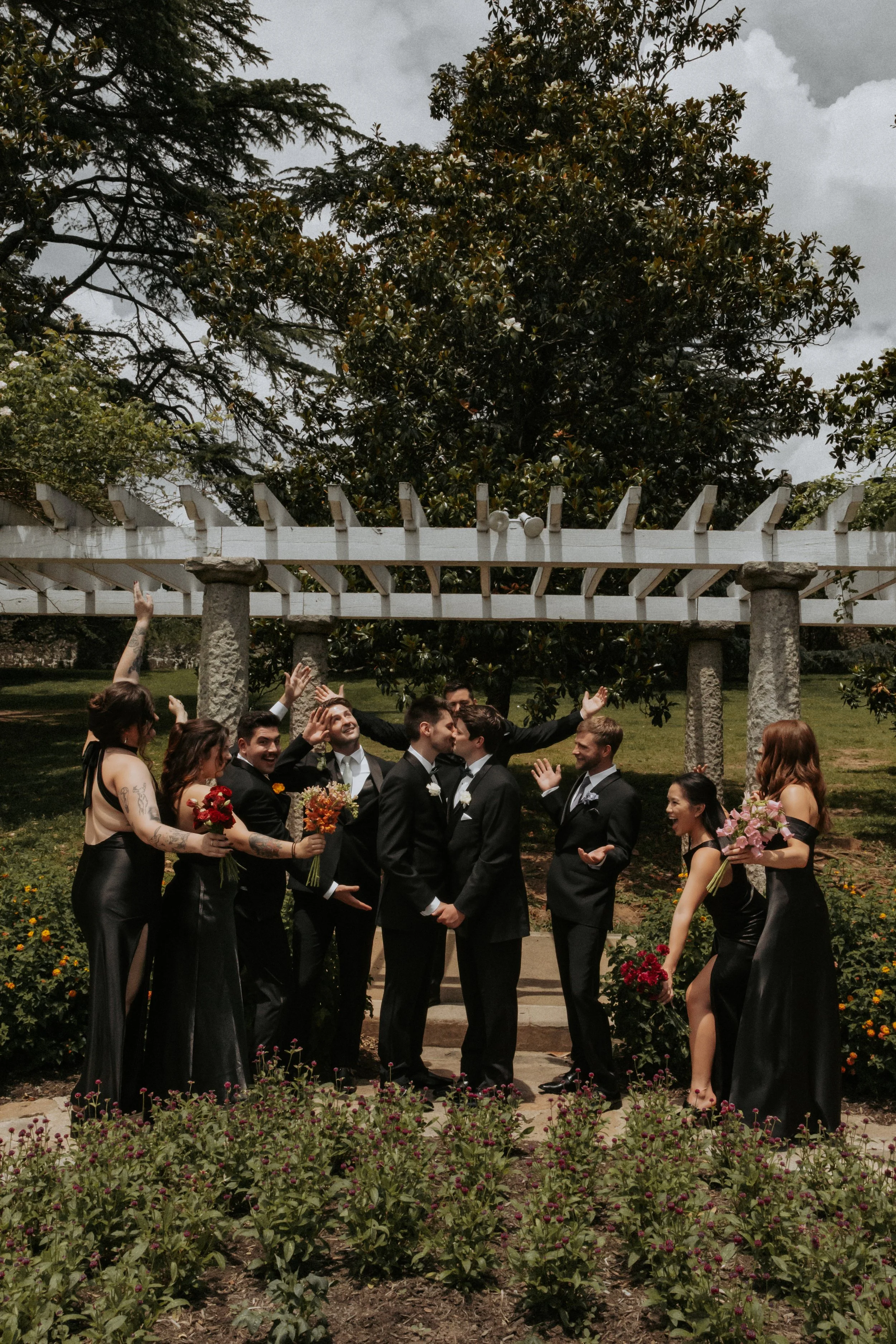 A wedding party outside, with the couple kissing in the center surrounded by bridesmaids and groomsmen, some holding bouquets, under a large tree and pergola.