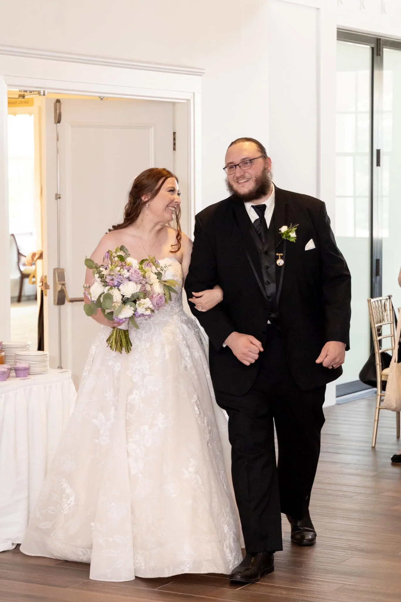A bride and groom walking arm in arm, smiling and looking at each other during a wedding reception inside a bright room with white walls and large windows.