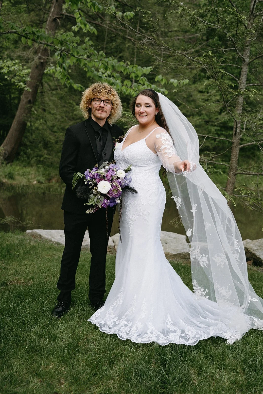 A newlywed couple standing outdoors near a river, with trees in the background. The bride is wearing a white lace wedding gown with a long train and veil, holding a bouquet of purple and white flowers. The groom is dressed in a black suit and tie.