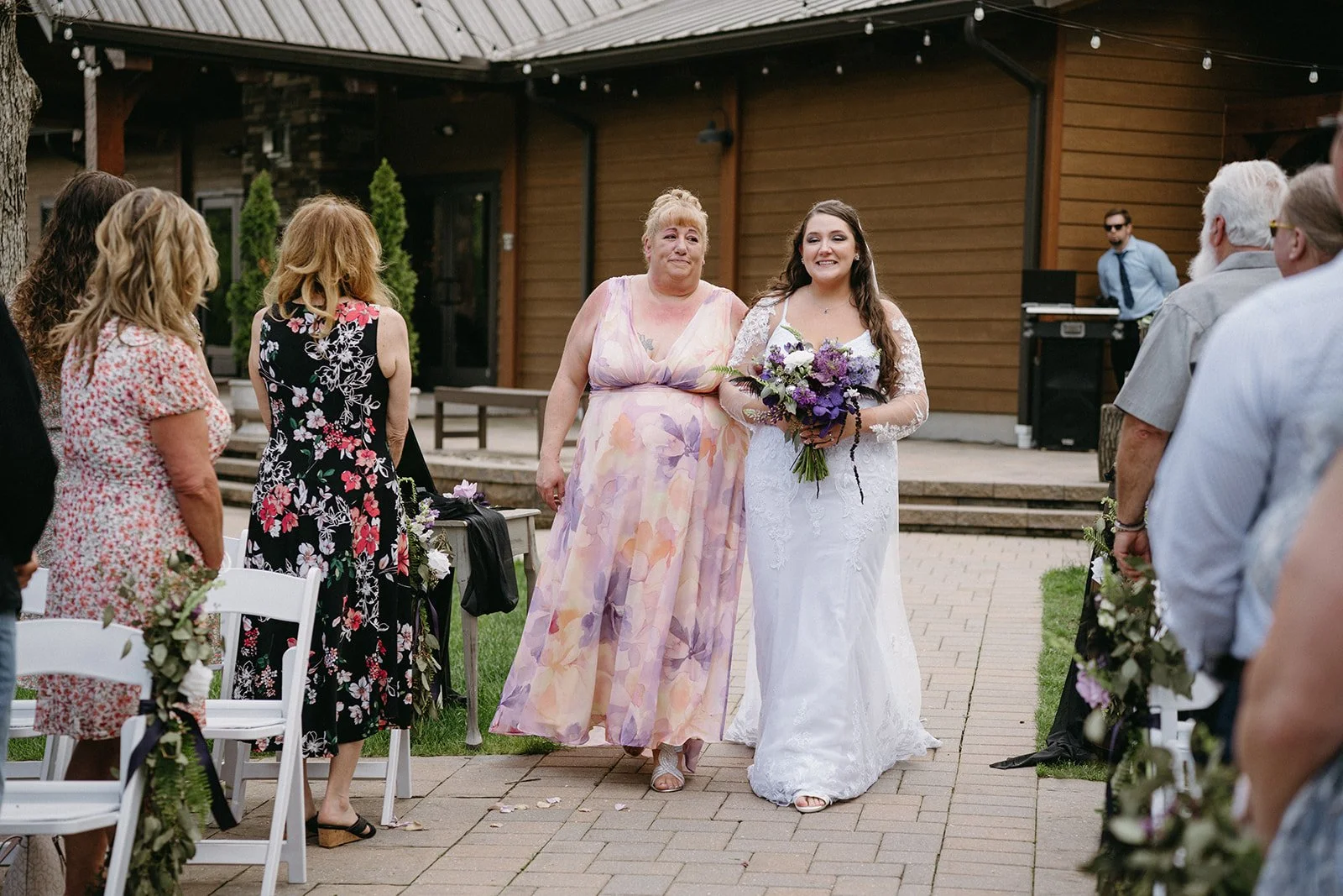 A bride in a white lace wedding dress holding a bouquet of purple flowers, walking down the aisle with a woman in a pastel floral dress at an outdoor wedding ceremony.