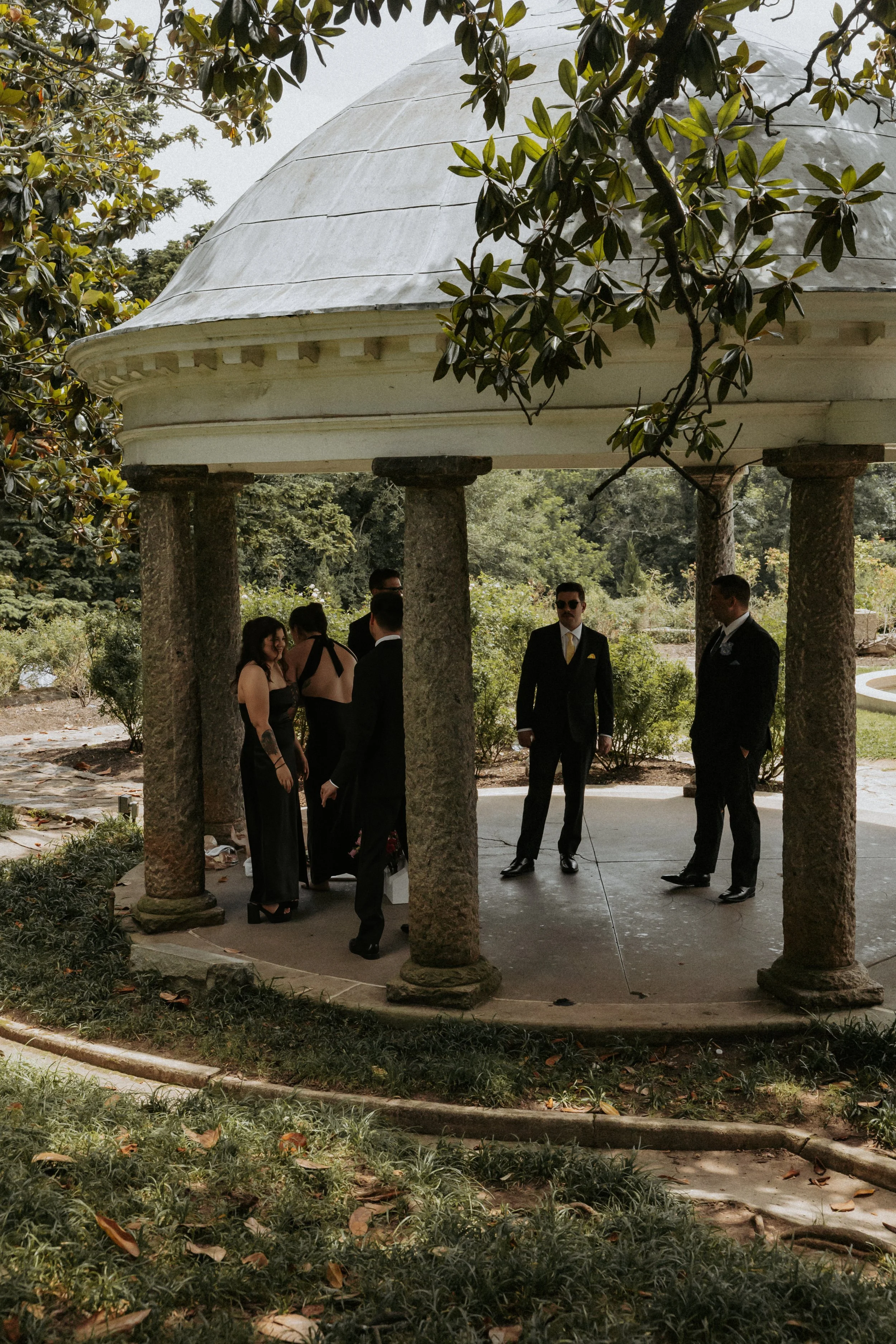 A group of people dressed formally, likely for a wedding, standing under a circular stone gazebo in a garden, surrounded by trees and greenery.