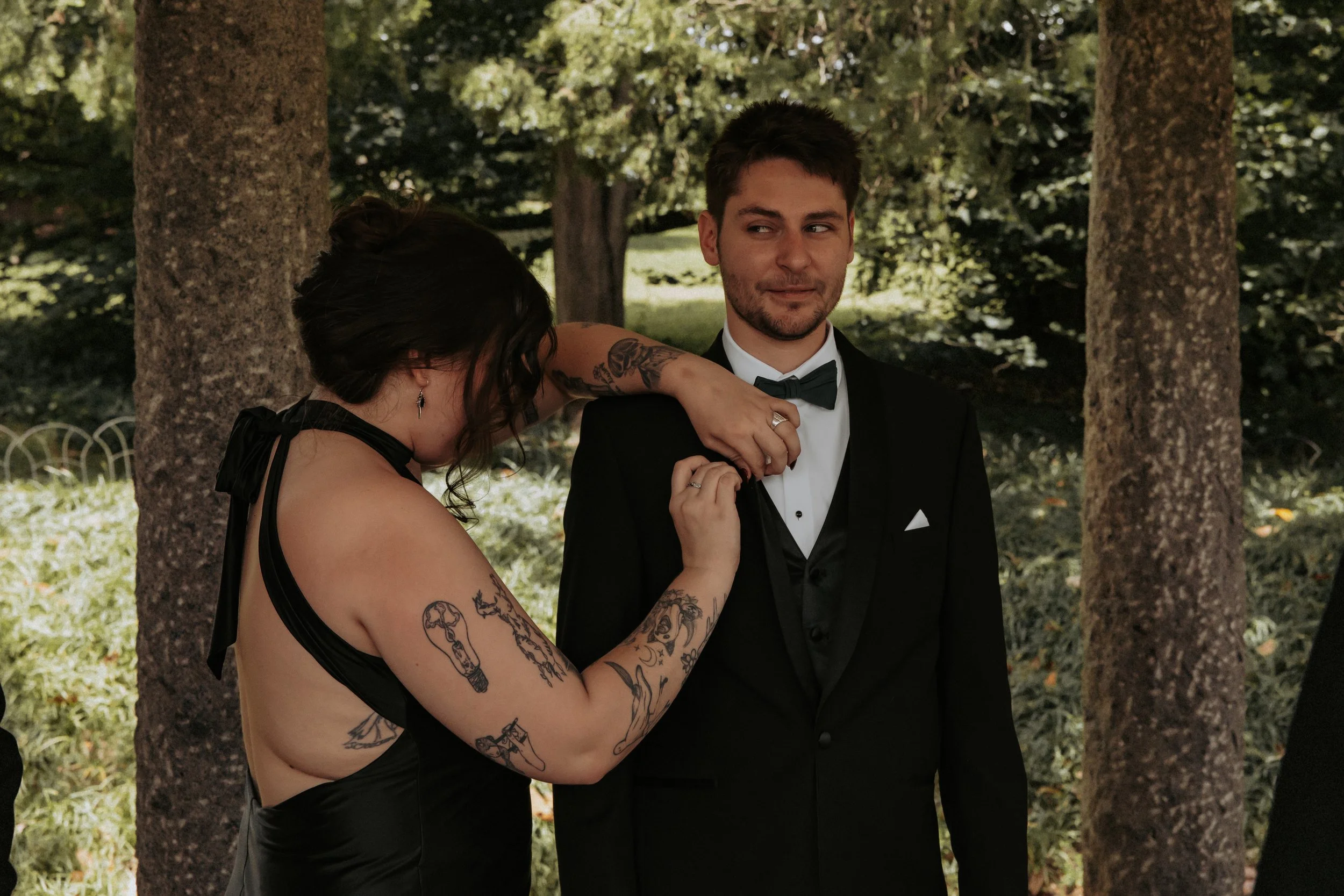 A woman helping a man adjust his tuxedo collar at an outdoor wedding or formal event, surrounded by trees.