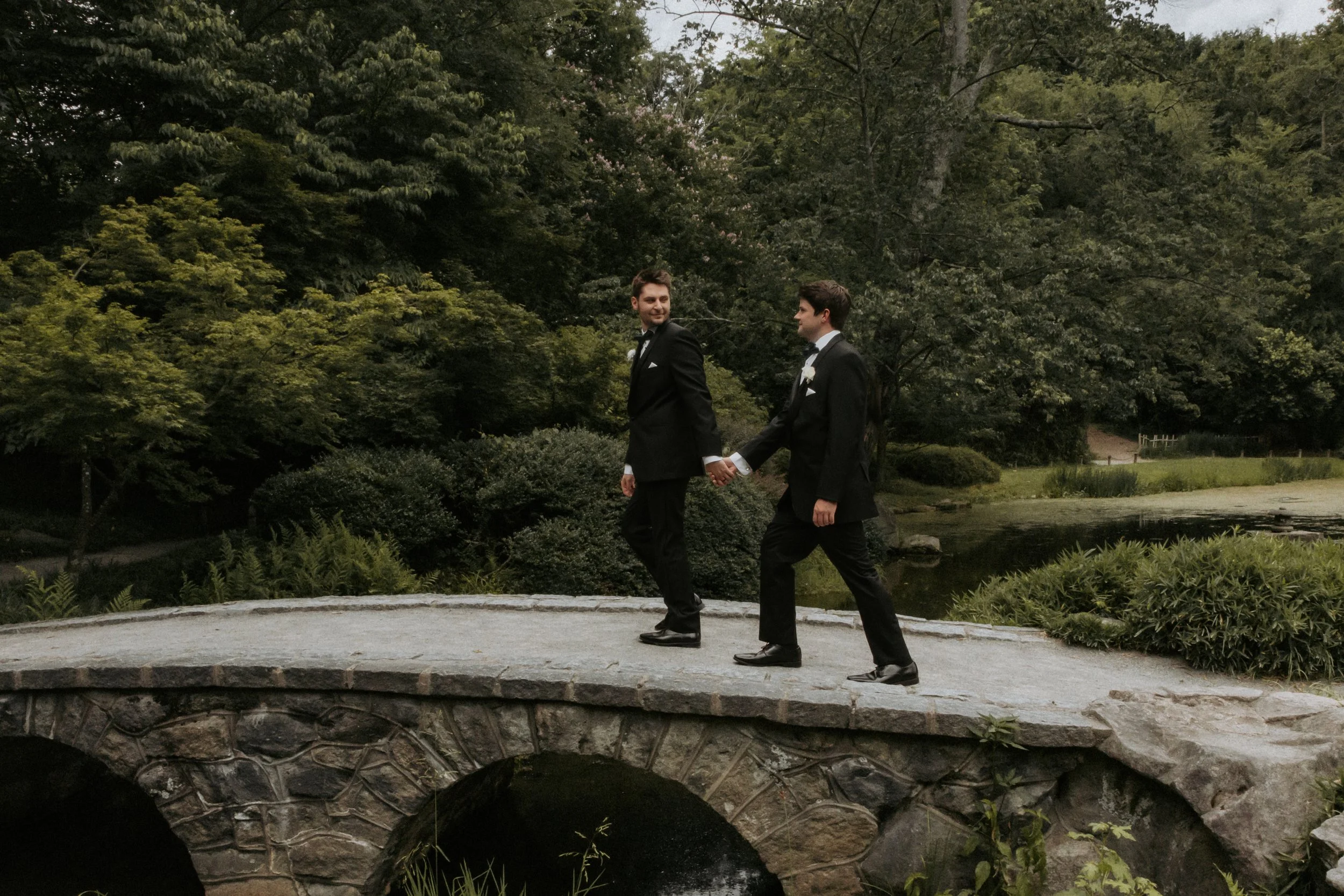Two men in tuxedos holding hands and walking on a stone bridge in a garden or park.