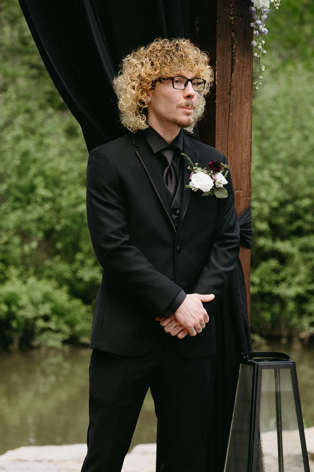 A man with curly blonde hair and glasses in a black suit stands outdoors, near a wooden post decorated with flowers, during a formal event.