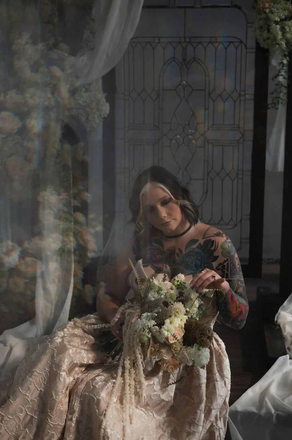 Woman with tattoos holding a bouquet of flowers, seen through a glass reflection, sitting in a vintage-style setting with stained glass window behind her.