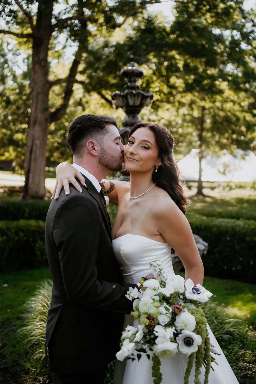 A newlywed couple embraces outdoors, with the groom kissing the bride's cheek, the bride holding a bouquet of white flowers, and a fountain and trees in the background.