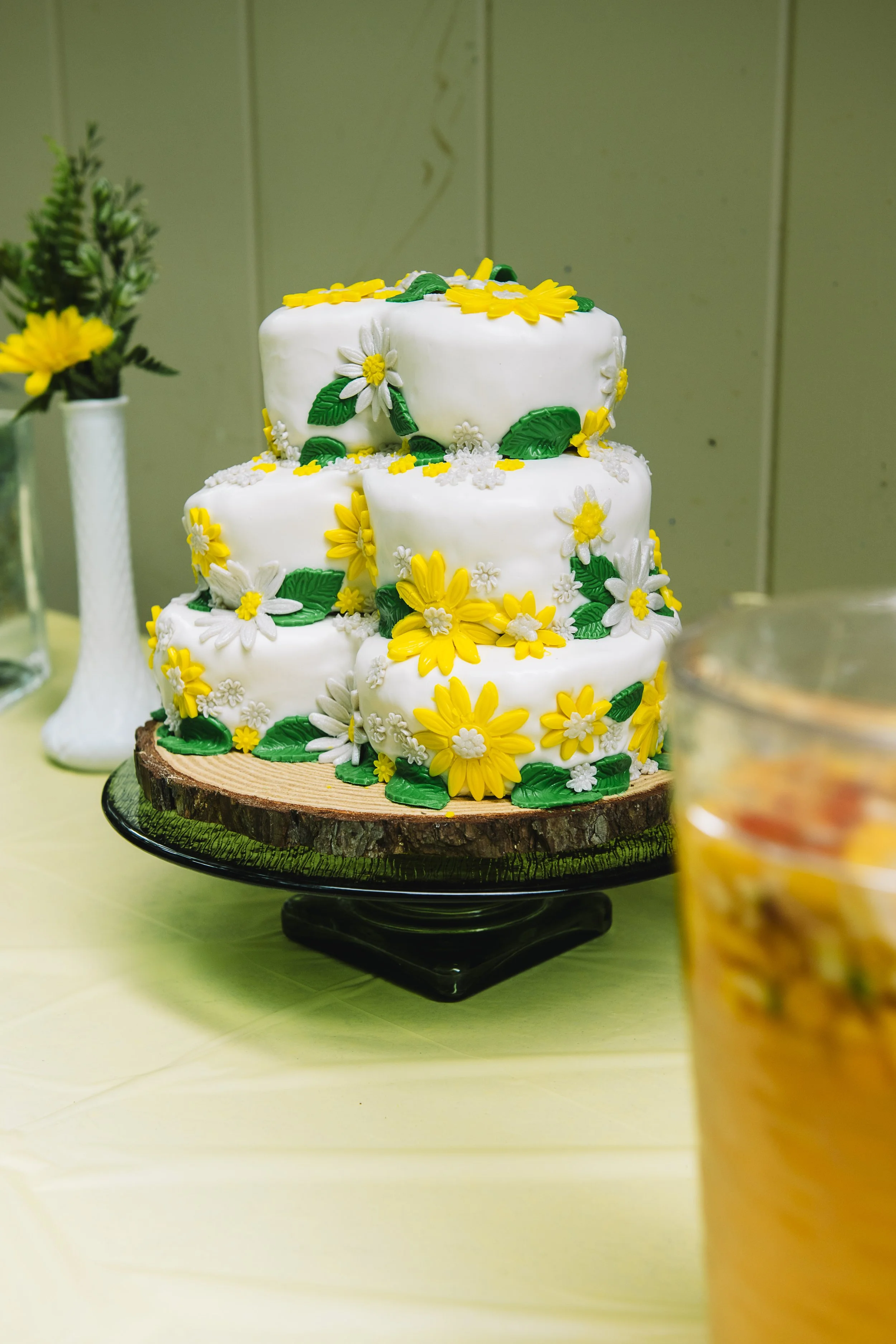 Three-tiered white cake decorated with yellow and white flowers and green leaves, displayed on a wood slice stand.