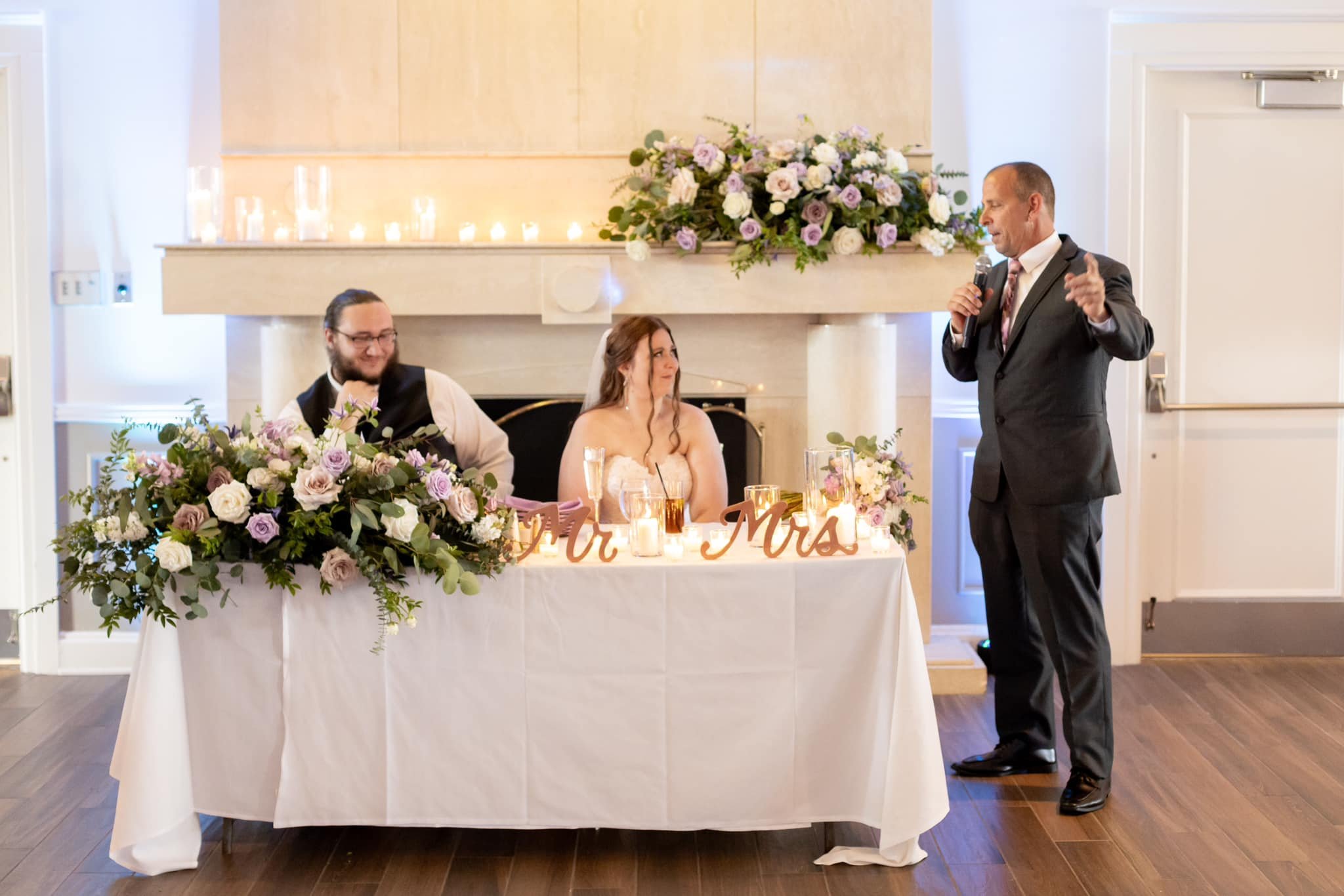 A wedding reception with a bride, groom, and another person sitting at a table decorated with flowers and candles. A man in a suit is standing and giving a speech.