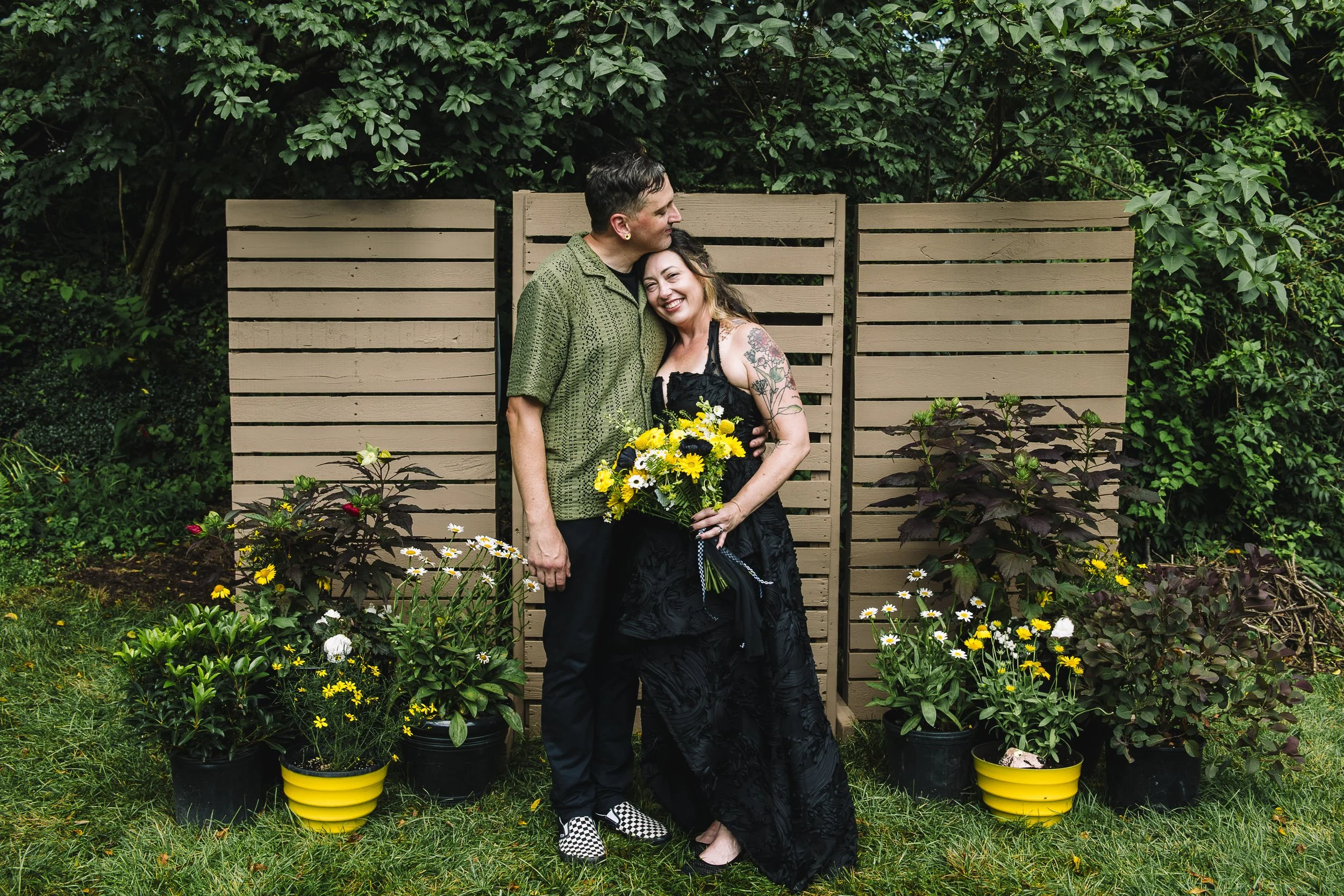 A couple standing in a garden, embracing each other, with the woman holding a yellow and white flower bouquet, surrounded by potted plants and a wooden fence.