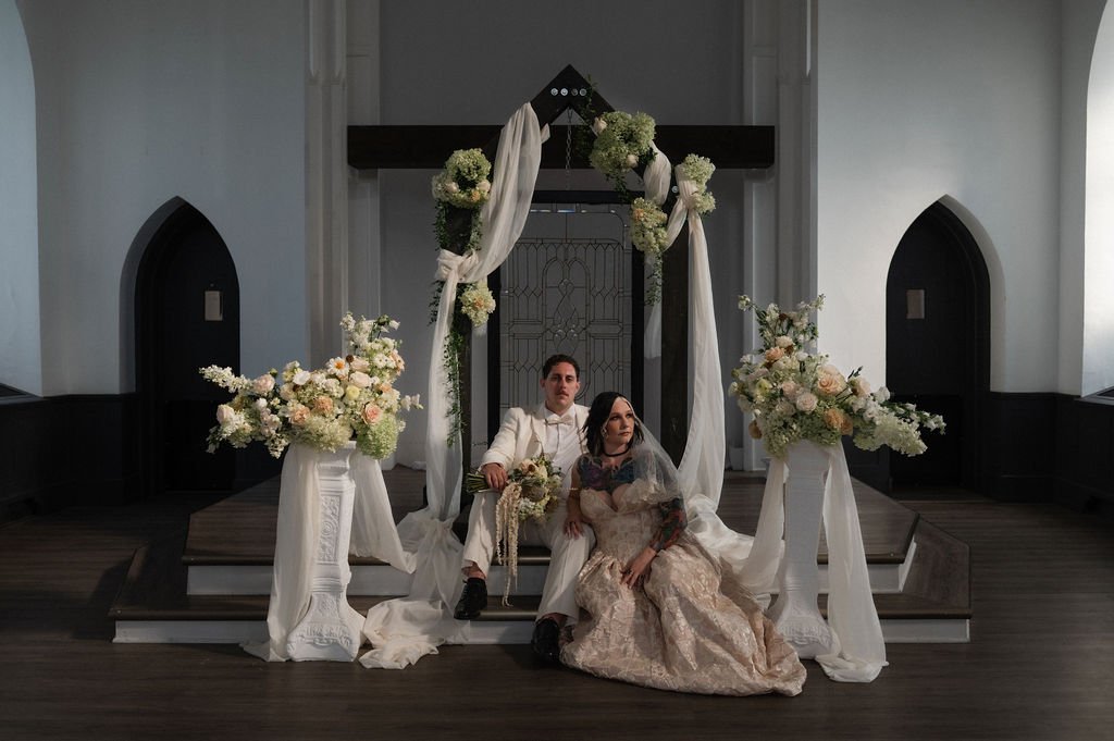A bride and groom sitting on a platform with floral arrangements and draped fabric during a wedding ceremony in a church.