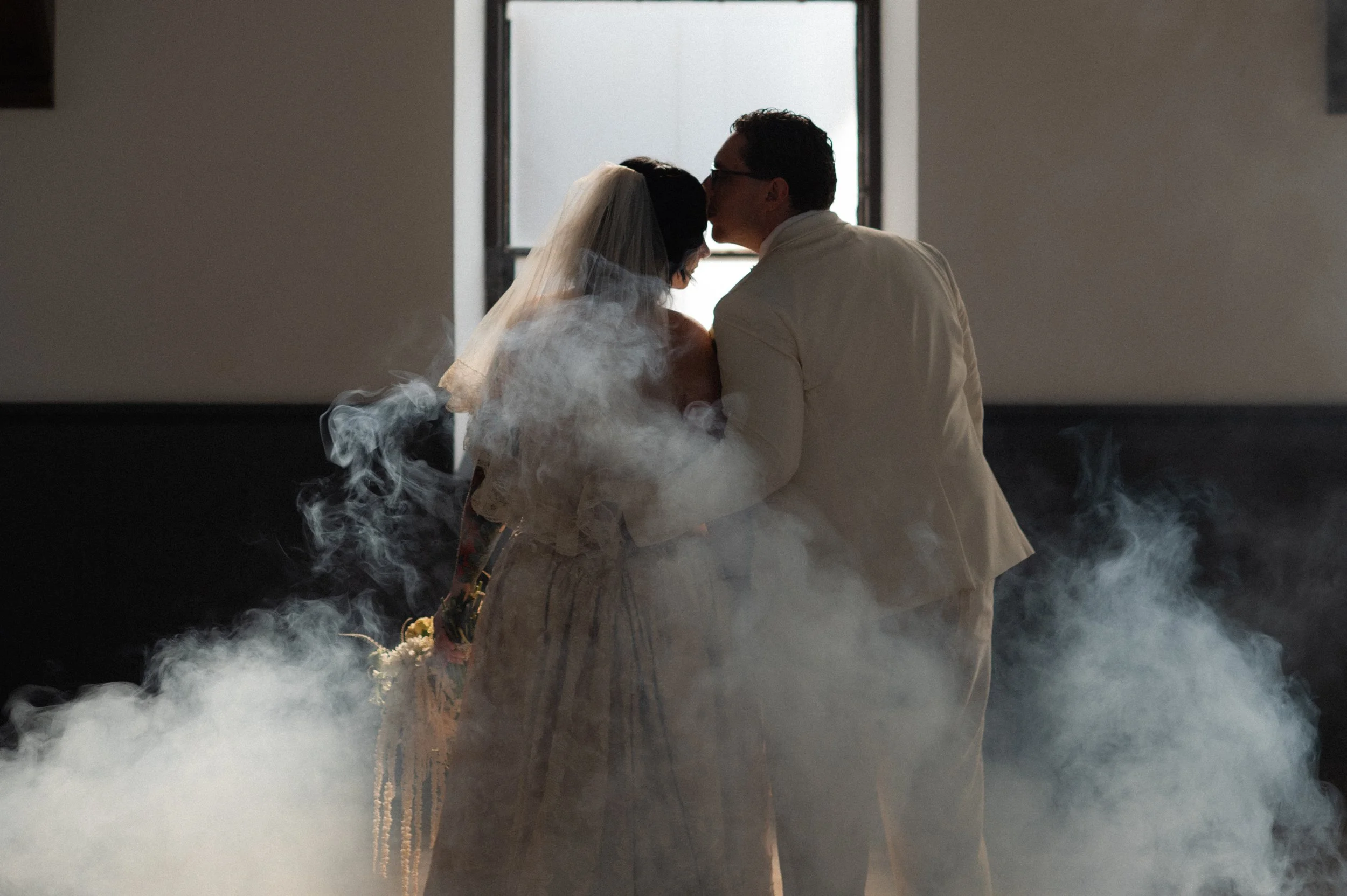 Couple, dressed in vintage wedding attire, sharing a tender moment with the groom kissing the bride's forehead. The scene is filled with white smoke or fog, and natural light is coming through an open window behind them.