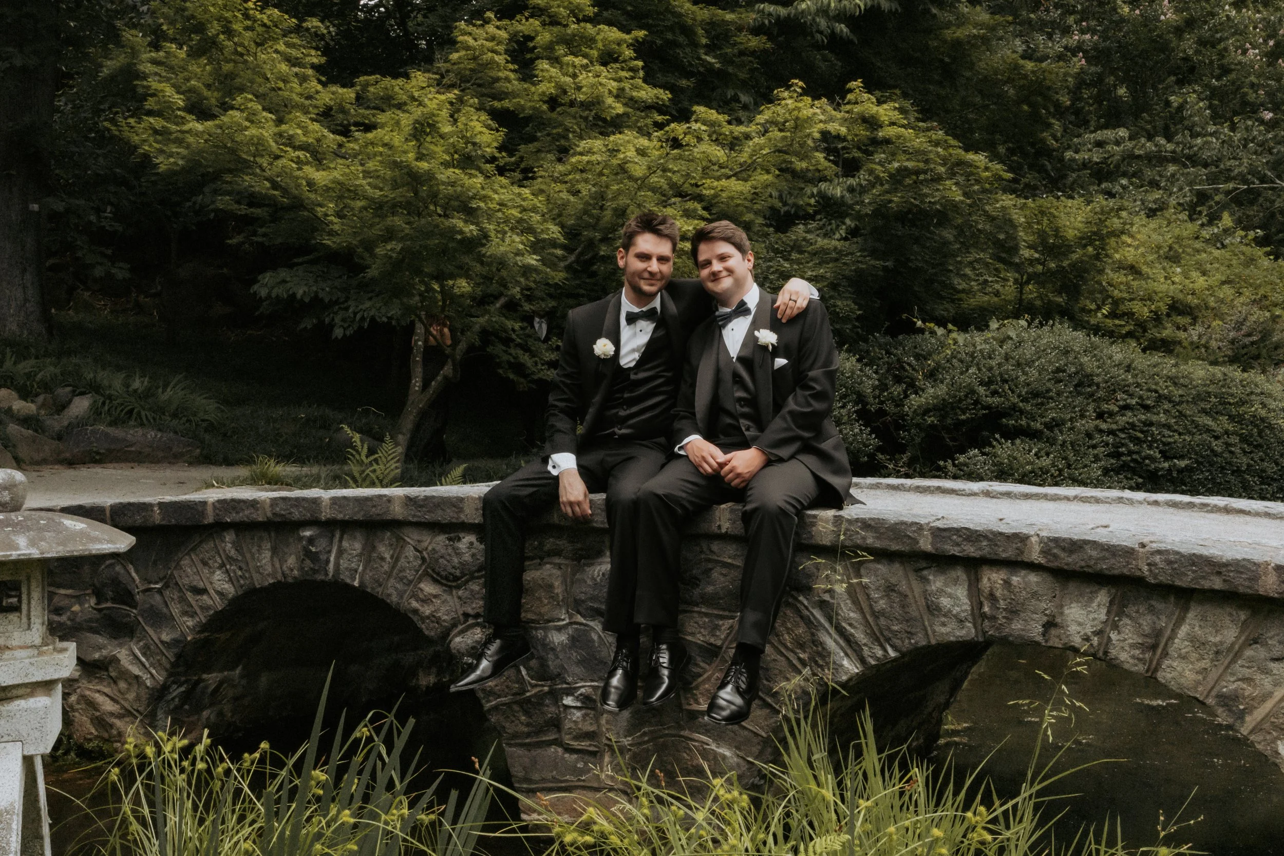 Two men in tuxedos sitting on a stone bridge in a lush outdoor garden, smiling and posing for a photo.