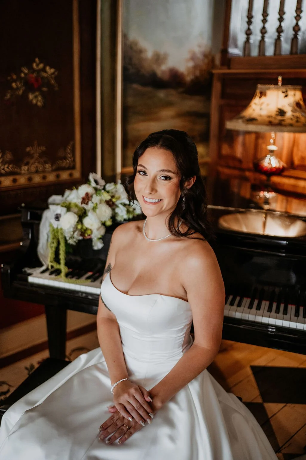 A woman in a wedding dress sitting at a piano with a floral arrangement on top, in a wood-paneled room with warm lighting.