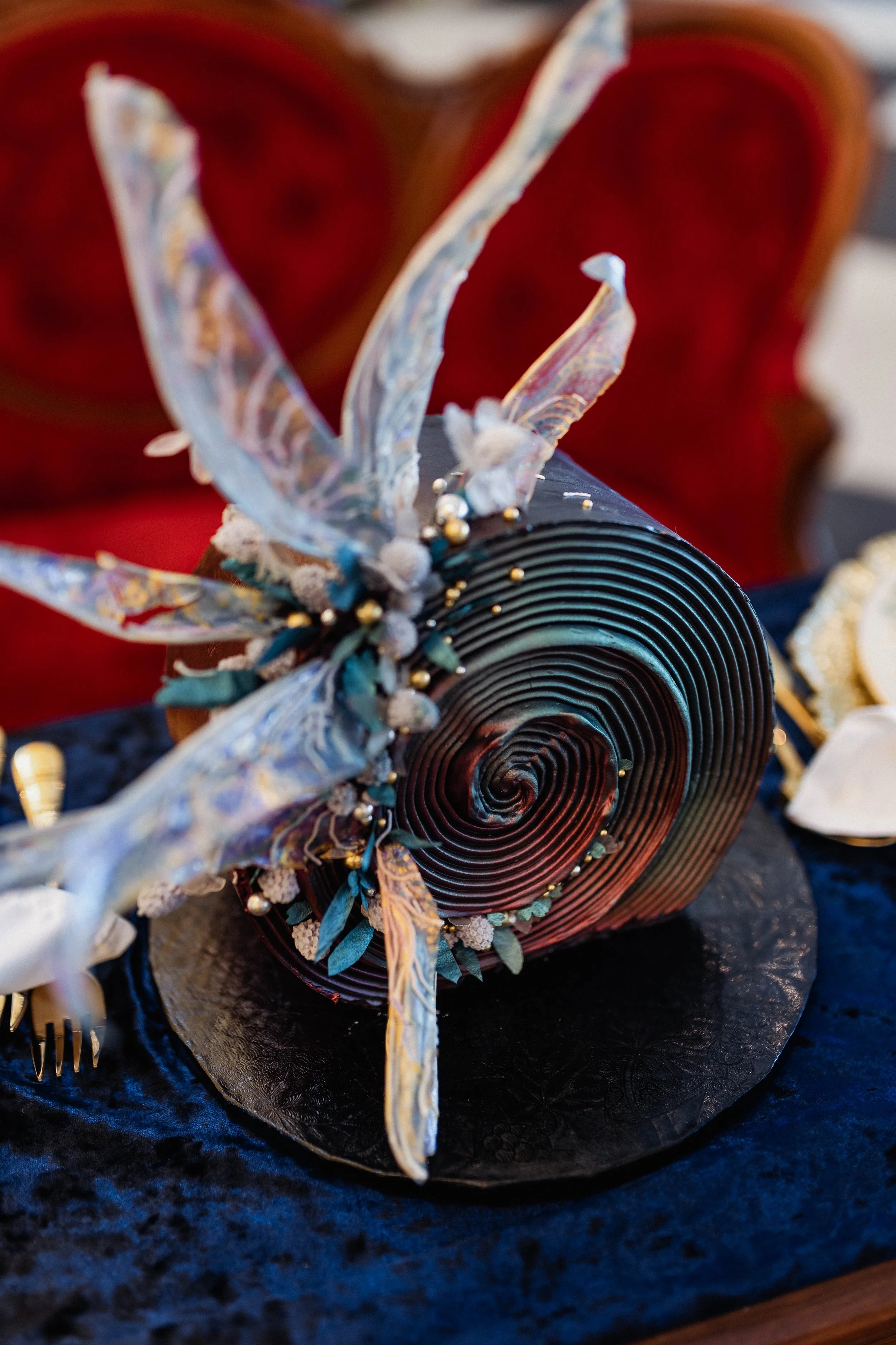 Decorative rolled black paper with intricate spiral design, adorned with fabric and pearl embellishments resembling a butterfly, placed on a dark blue cloth.