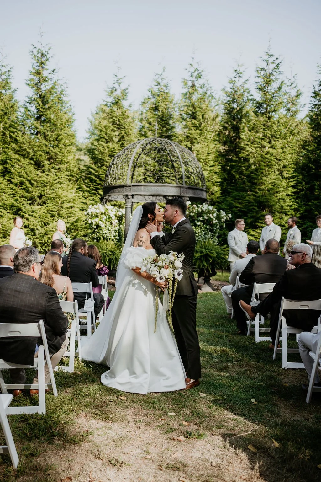 A bride and groom kissing at their outdoor wedding ceremony surrounded by guests and lush greenery.