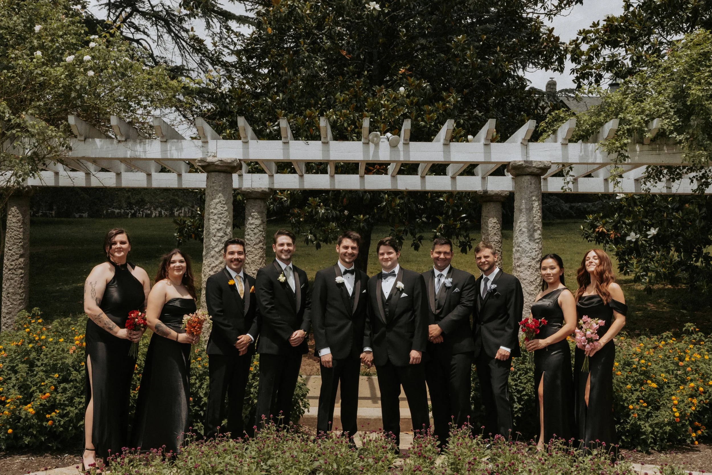 Group of eleven people in formal attire posing outdoors, with a garden and white wooden trellis behind them.
