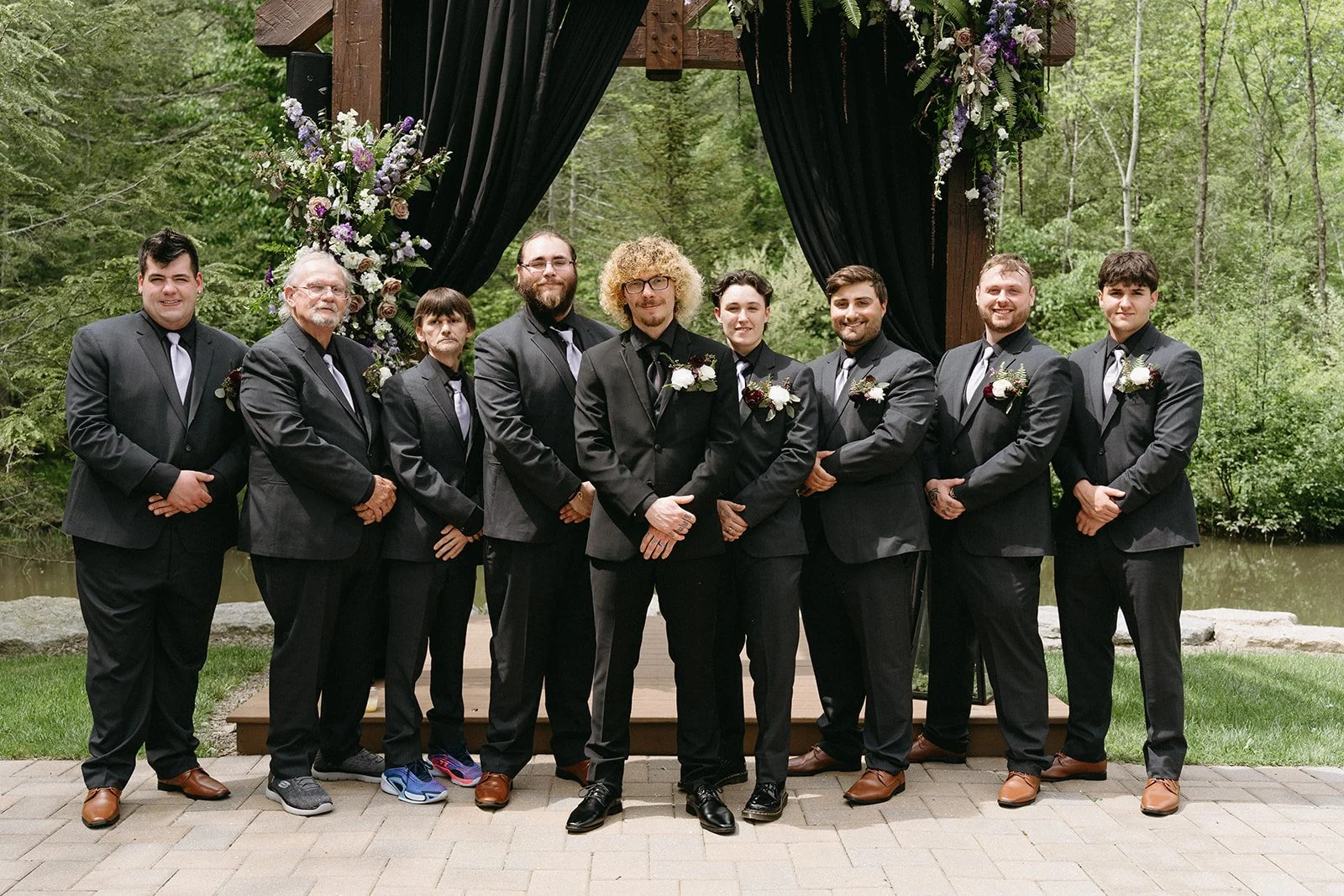 Group of nine men dressed in dark suits with ties standing together outdoors at a wedding ceremony, posing in front of a decorated wooden arch with flowers and black drapes, with green trees and grass in the background.
