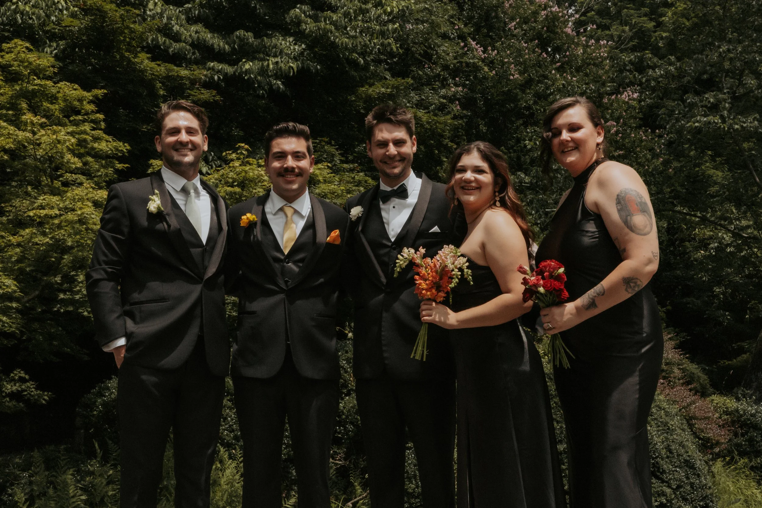 Group of six people in formal attire, standing outdoors in front of green foliage and trees, smiling at the camera.