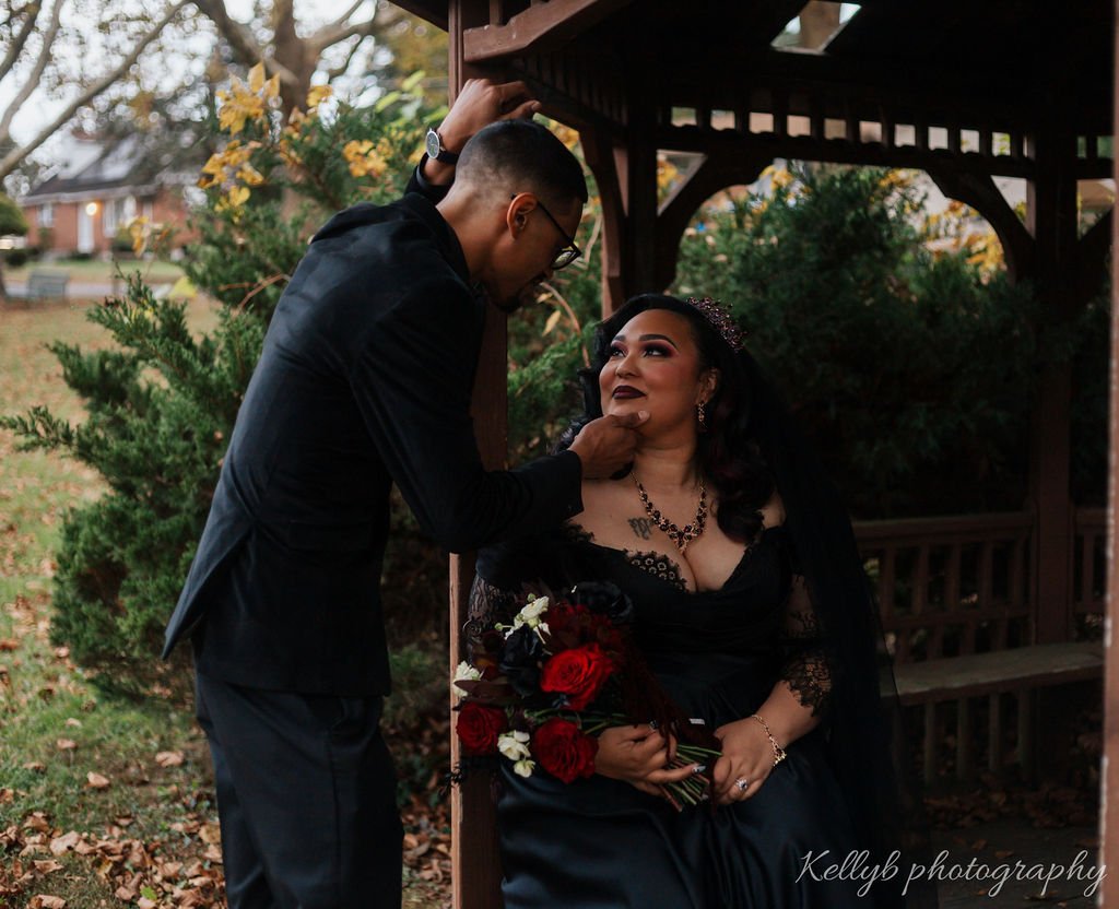 A man dressed in a black suit is touching up a woman's makeup in a park, with autumn trees and houses in the background. The woman, dressed in black with jewelry and a tiara, sits holding a bouquet of red roses.