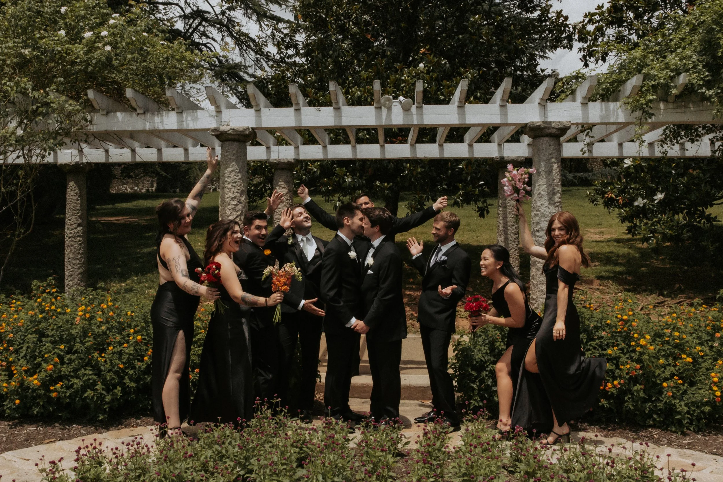 A group of wedding party members, dressed in formal black attire, standing outdoors underneath a white trellis with stone pillars, surrounded by flowers and greenery, celebrating and sharing a kiss.