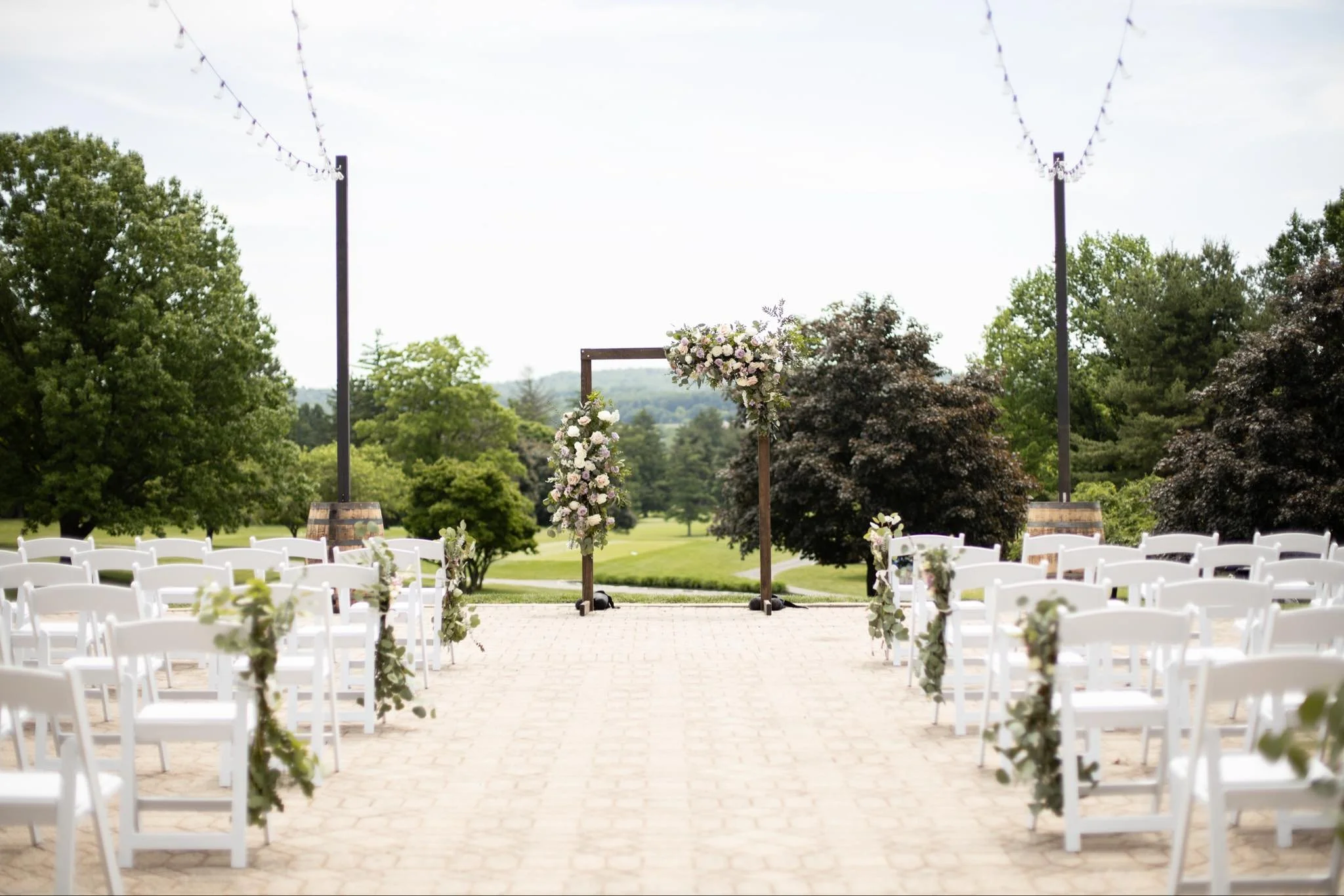 Outdoor wedding ceremony setup with white chairs, floral decorations, and an arch over a paved pathway, surrounded by trees and greenery.