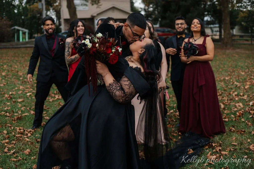 A couple shares a romantic kiss during their wedding outdoors, with friends in formal attire smiling in the background, surrounded by fallen autumn leaves.