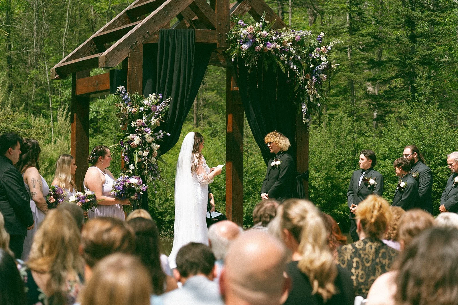 Outdoor wedding ceremony with bride, groom, officiant, bridesmaids, and groomsmen under a wooden arch decorated with purple and white flowers, set in a lush green forest.