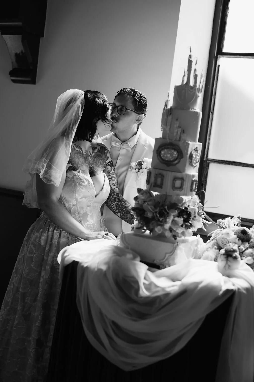 Black and white photo of a newlywed couple kissing in front of a wedding cake, with flowers on the table and a window in the background.
