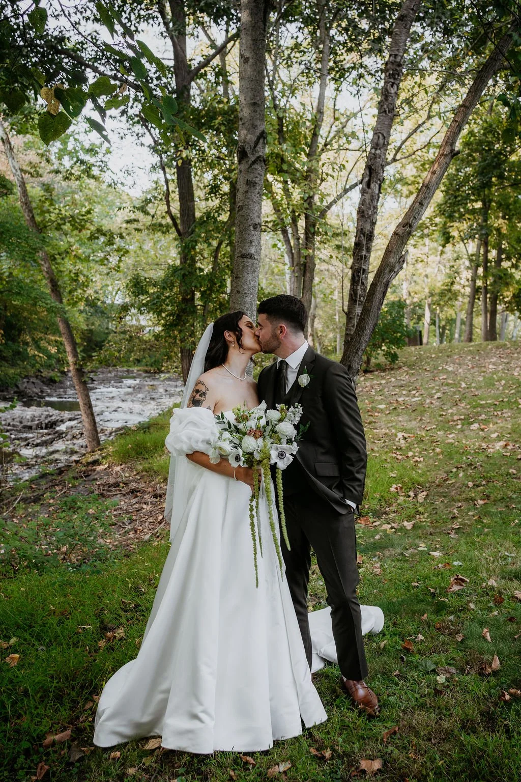 A bride and groom kiss outdoors in a wooded area, with the bride holding a bouquet of white and green flowers, and both dressed in formal wedding attire.