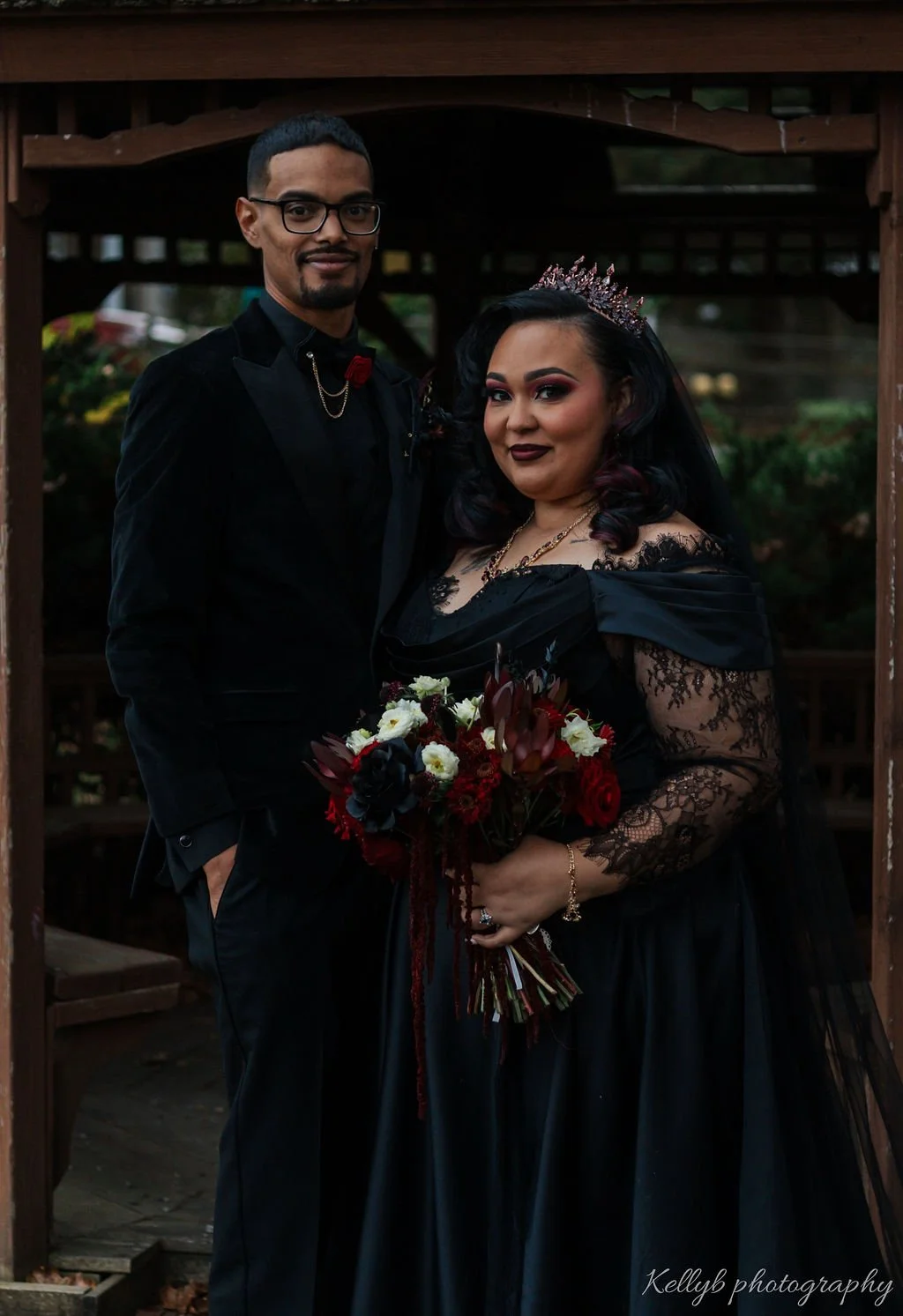 A couple dressed in black, with the woman wearing a crown and holding a bouquet, standing under a wooden structure.