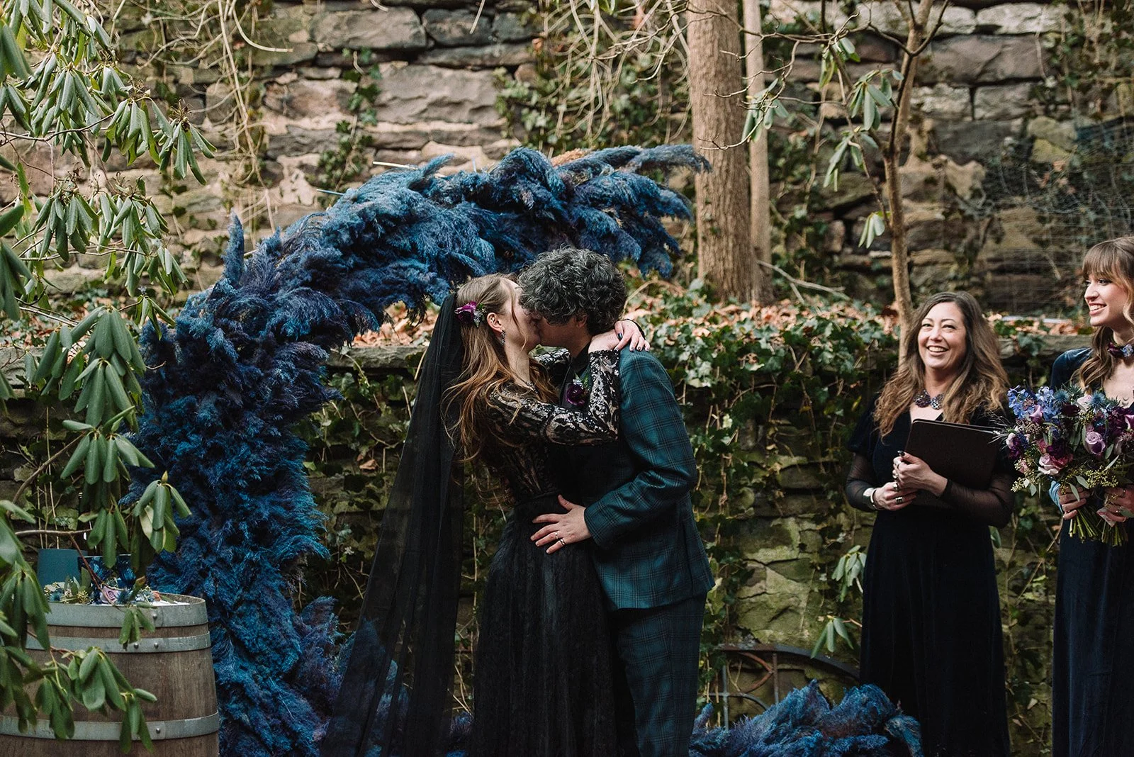 A couple kissing during a wedding ceremony outdoors, with two women standing nearby. One woman holds a bouquet, and another holds a clipboard, both smiling. The ceremony is set against a stone wall with plants.