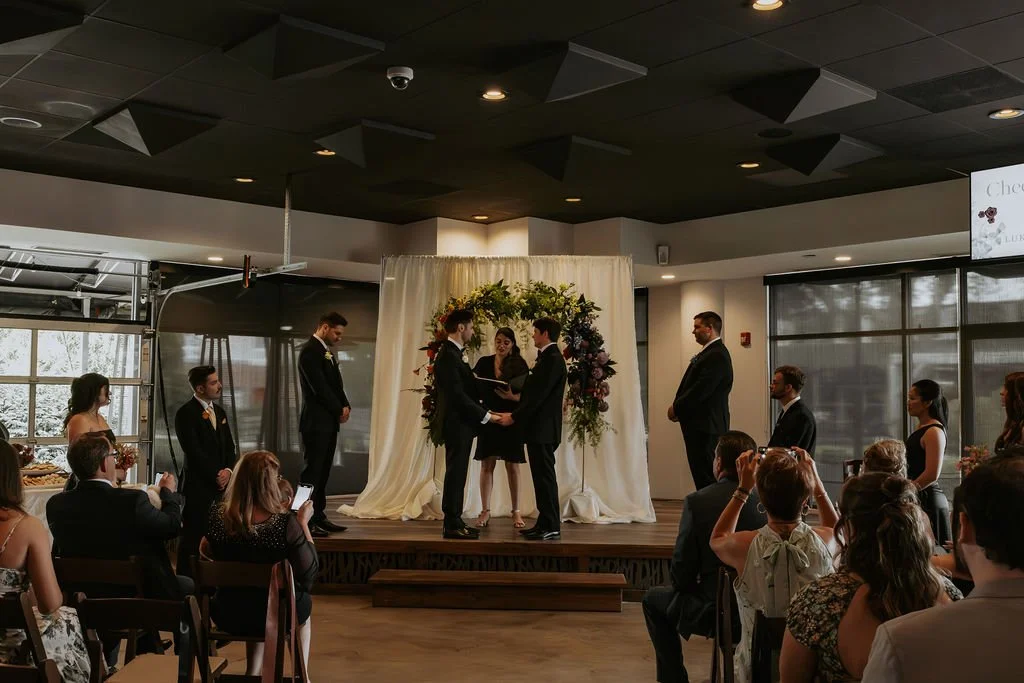 A wedding ceremony on a small stage with a couple exchanging vows, officiated by a woman, surrounded by friends and family in a modern indoor venue.