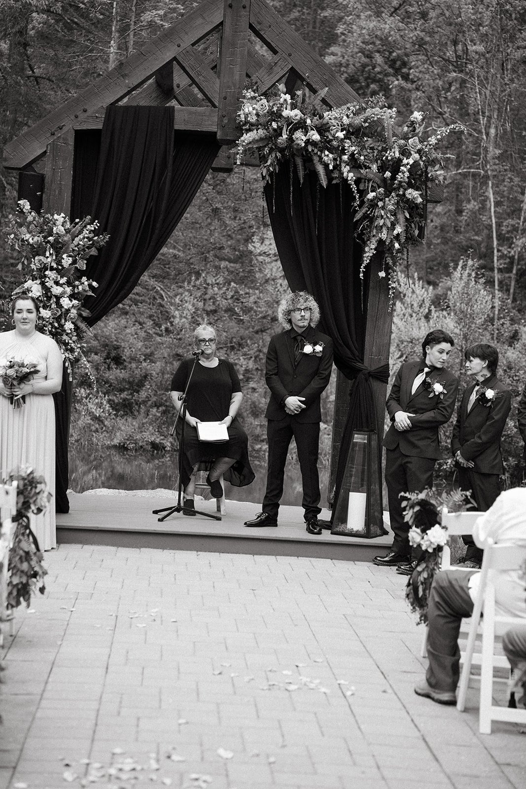 Black and white photo of an outdoor wedding ceremony with a wooden arch decorated with flowers and black curtains, surrounded by trees and guests.