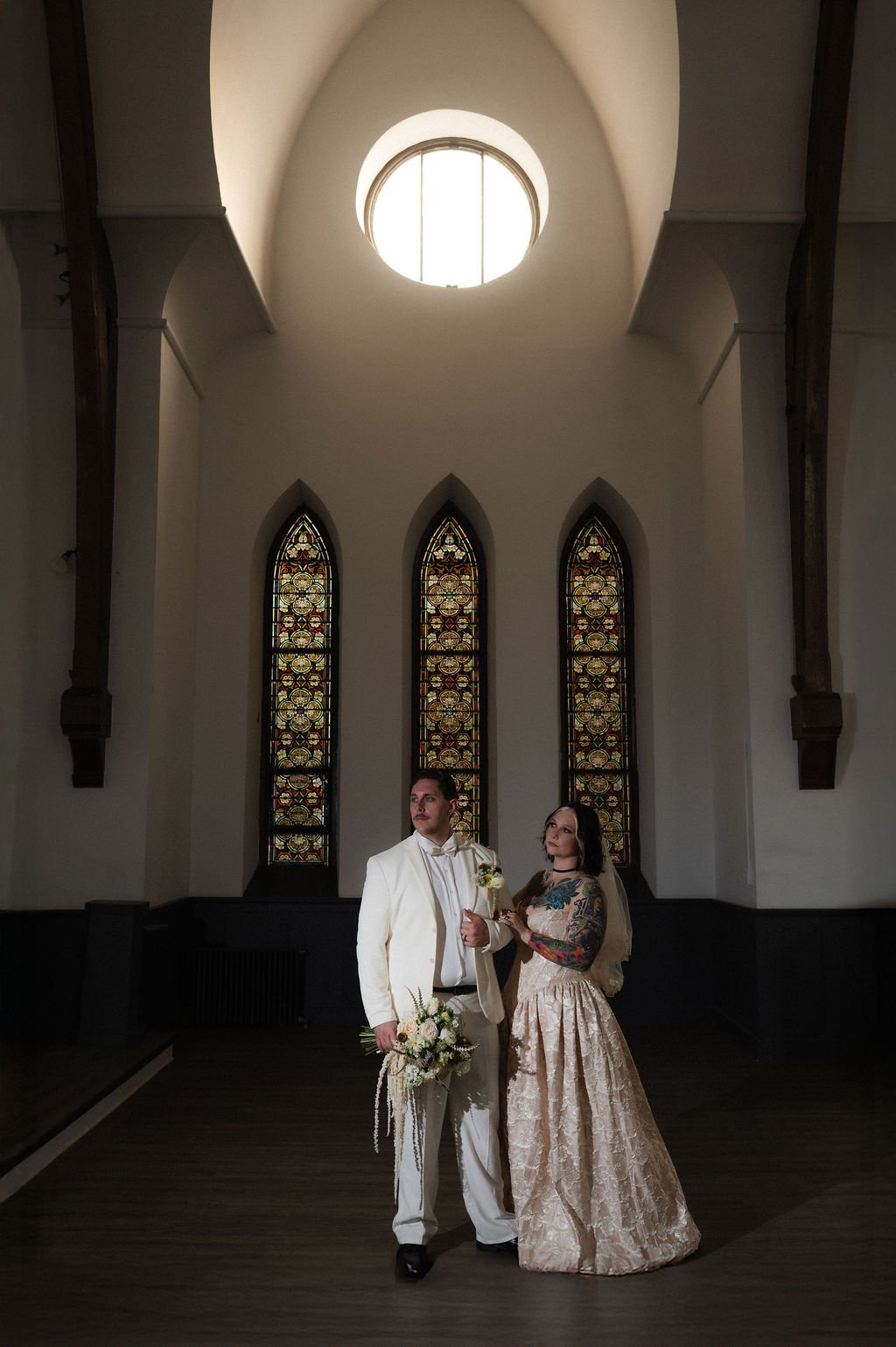 A couple dressed in wedding attire standing inside a church with tall stained glass windows and a high vaulted ceiling with a round window at the top.