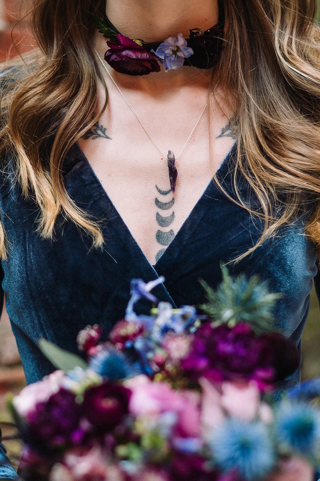 A woman wearing a velvet dress, a floral necklace, and a moon phase tattoo on her chest, holding a bouquet of flowers.