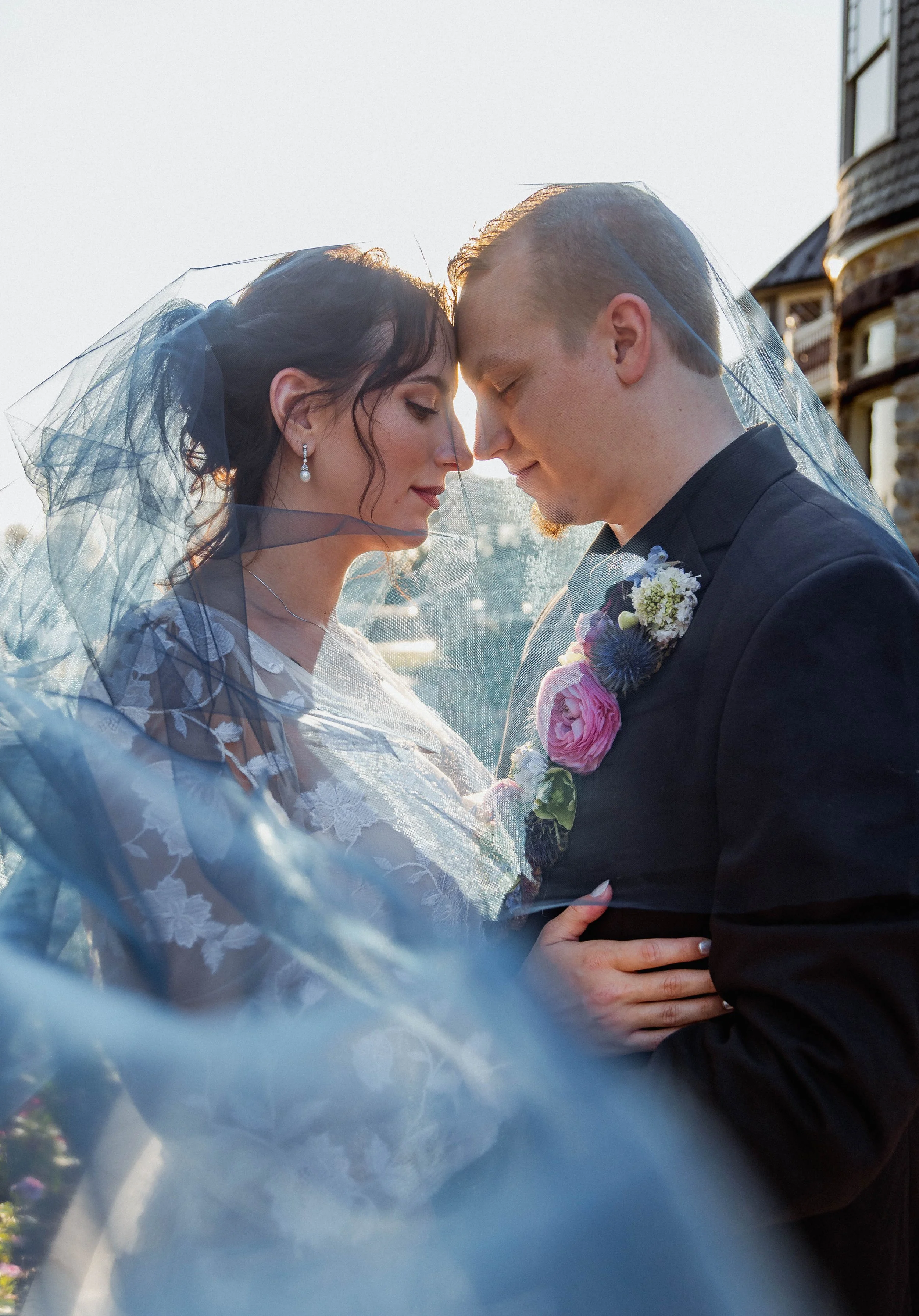 Bride and groom with foreheads touching, wrapped in wedding veil, outdoors during sunset, romantic moment.