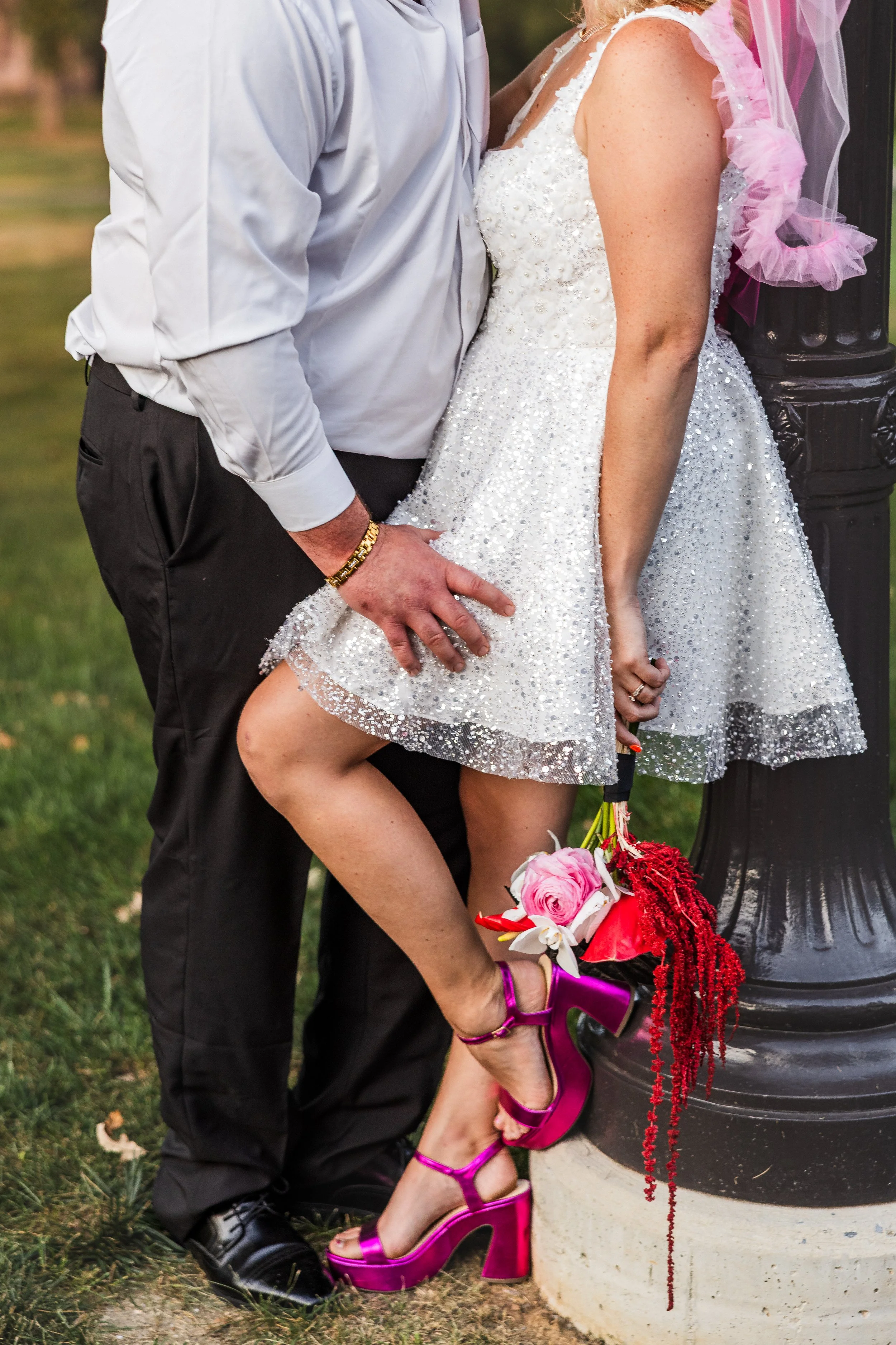 Close-up of a couple, with the man in a white dress shirt and black pants and the woman in a sparkling white dress, holding a bouquet of pink and red flowers, standing outdoors near a black lamppost.