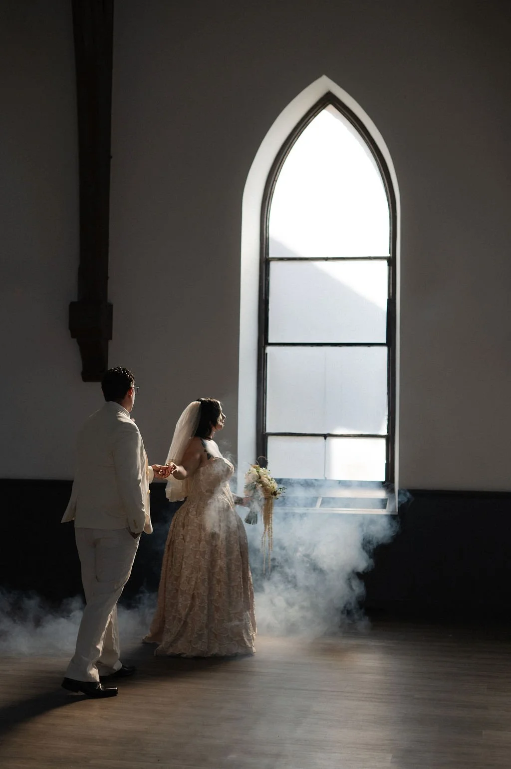 A bride and groom holding hands during their wedding ceremony in a church with a tall arched window and fog on the floor.