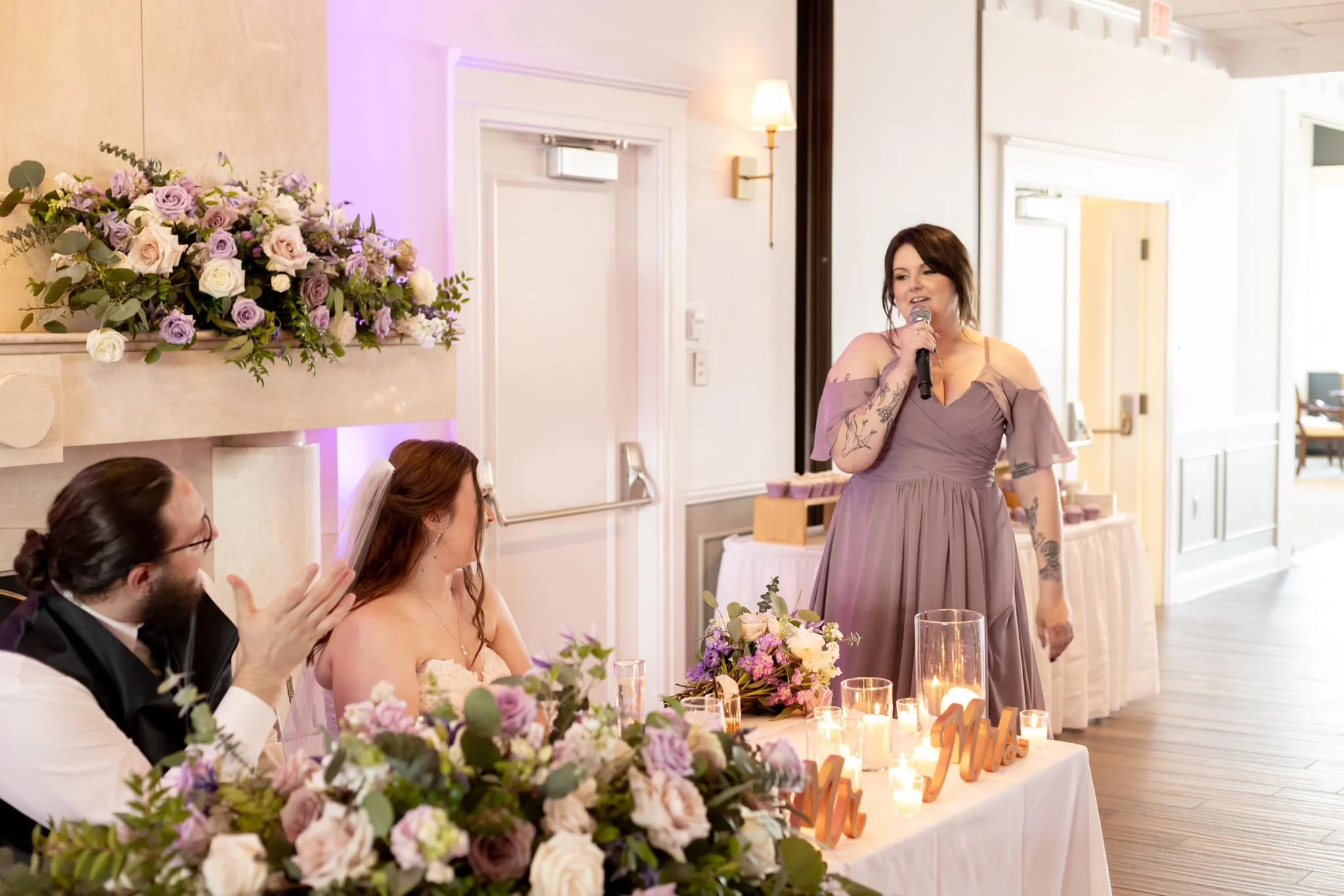 A woman giving a speech at a wedding reception, standing next to the bride and groom sitting at a decorated table with flowers and candles.