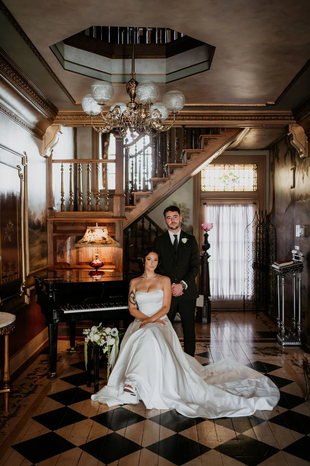 A bride and groom in formal wedding attire pose inside a vintage, elegantly decorated room with a grand staircase, chandelier, stained glass window, and a piano.