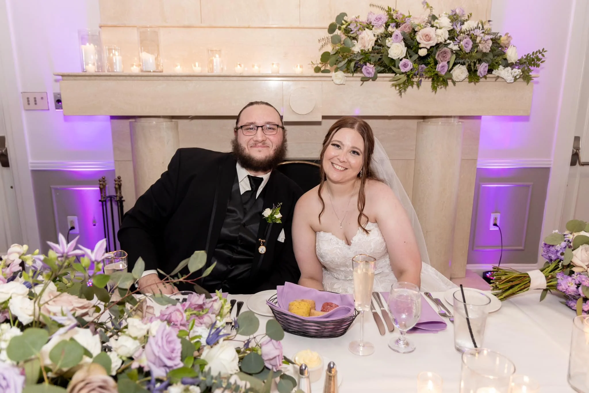 Bride and groom sitting at a wedding reception table, smiling, with floral arrangements and candles in the background.