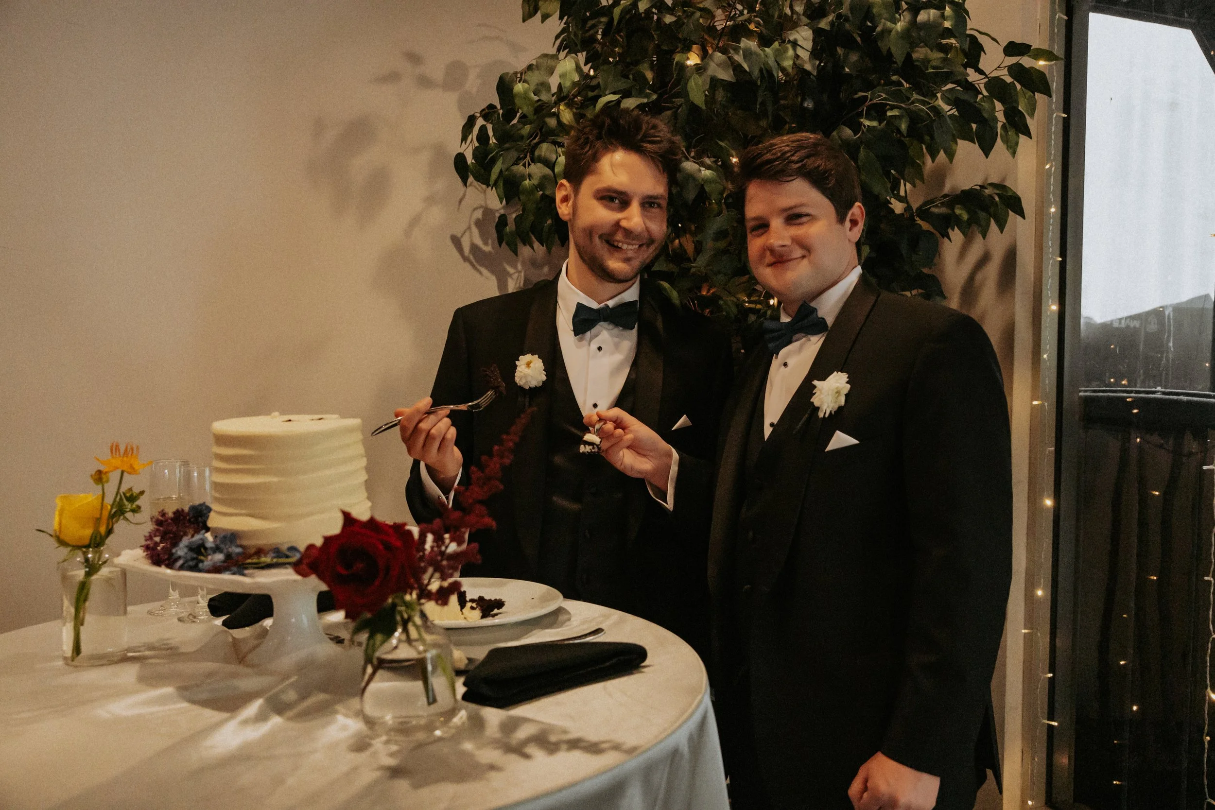 Two men in tuxedos with bow ties standing at a wedding reception, smiling and cutting the wedding cake together. There are flowers on the table and a large leafy plant in the background.