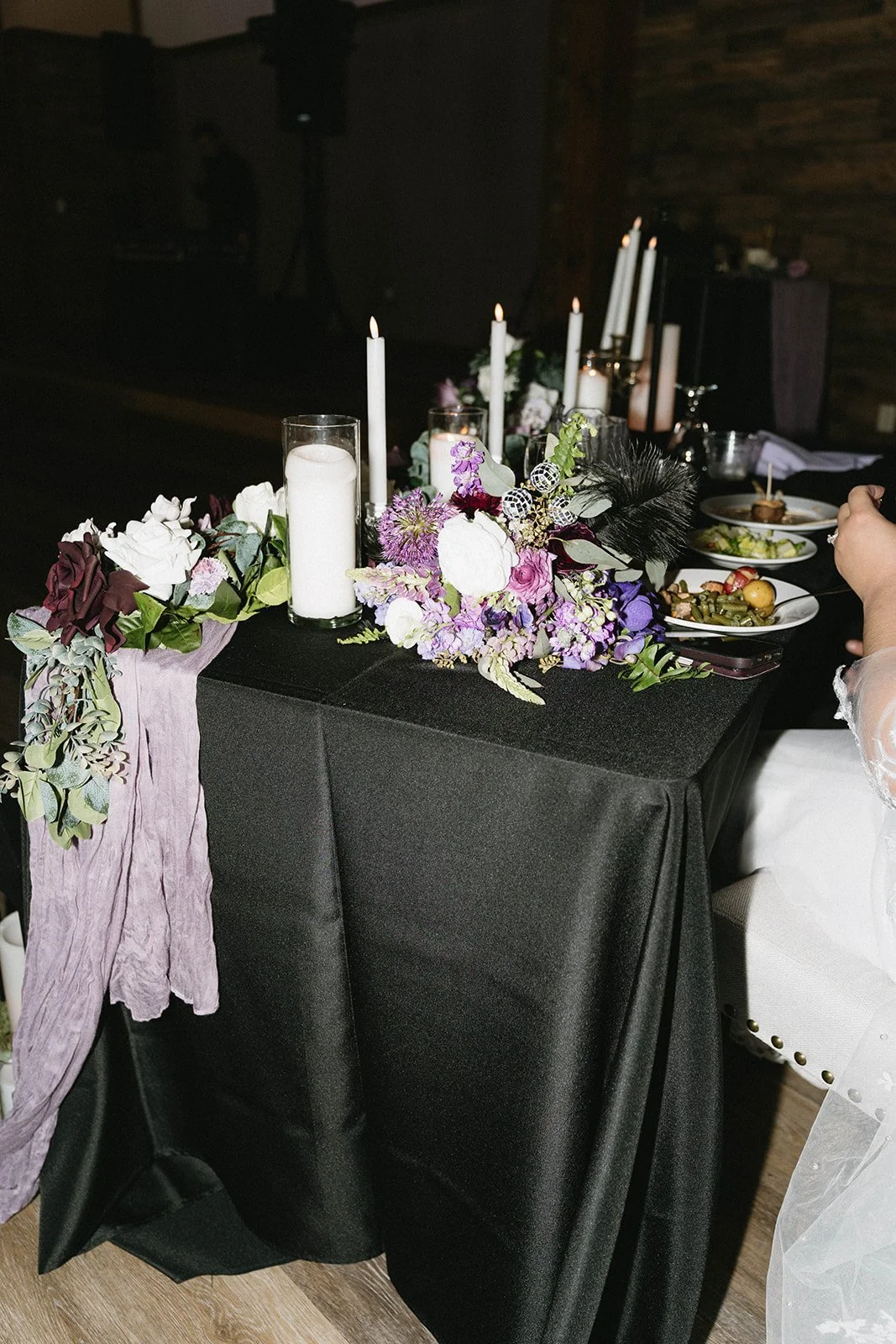 A table decorated with white candles, purple, white, and green floral arrangements, and a purple fabric drape at a formal event.