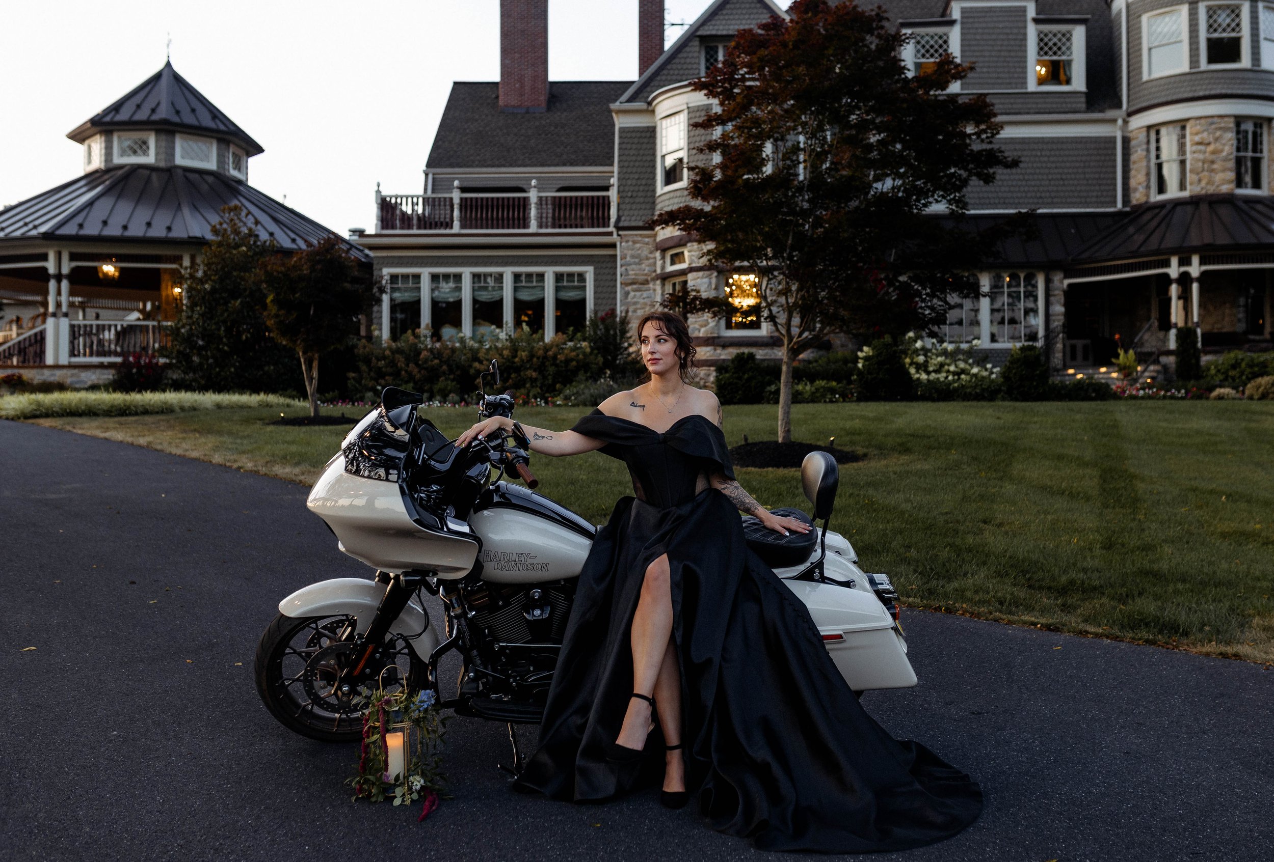 Woman in black formal dress sitting on a white Harley-Davidson motorcycle in front of a large house at dusk.